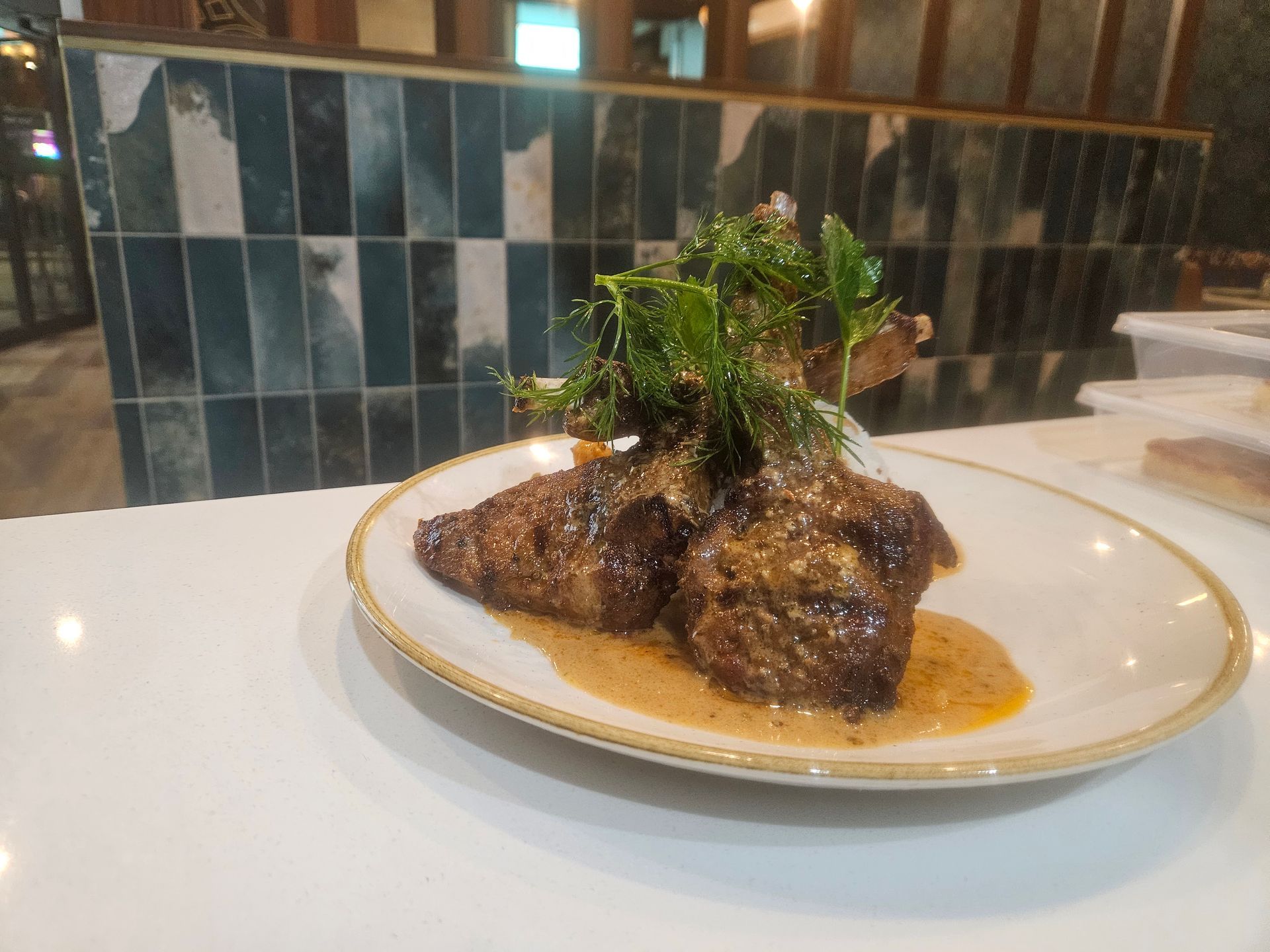 Lamb chops on a white plate, topped with greens, sitting on a white surface with a tiled background.