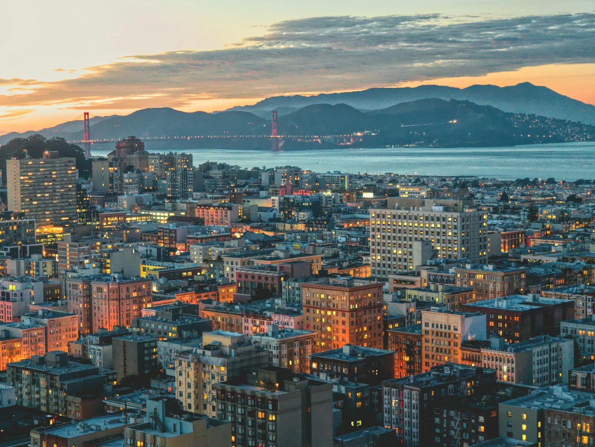 Dusk cityscape with glowing buildings, bay, and a distant bridge under a cloudy sunset sky