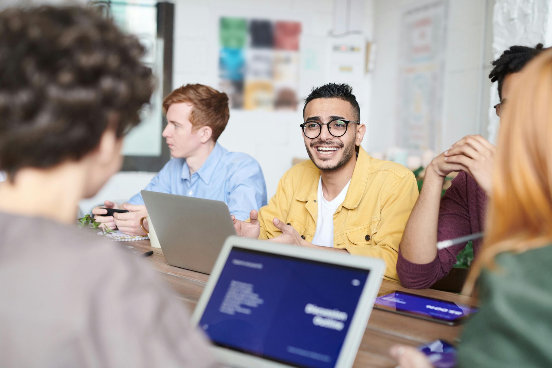 People gathered around laptops in a bright meeting room, discussing work.