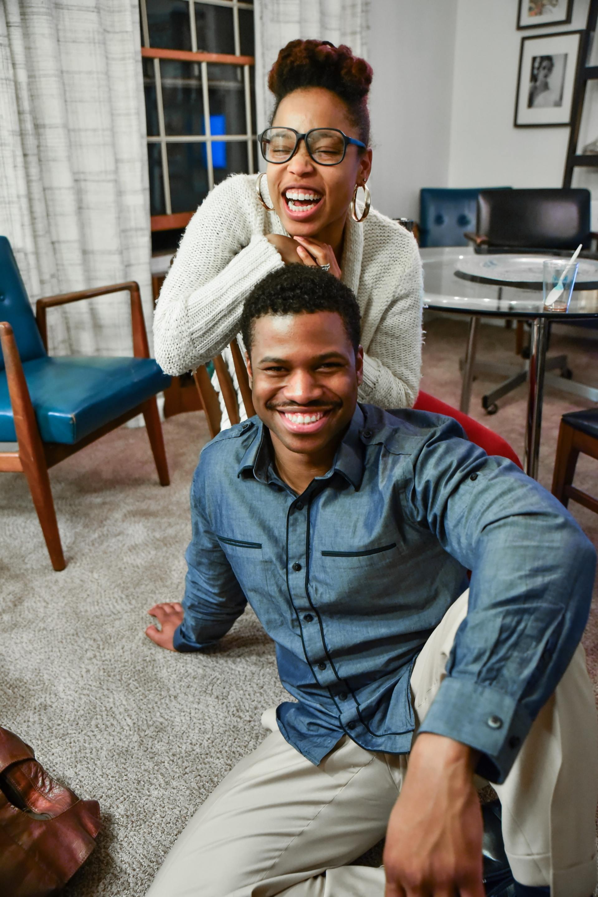 Woman smiles over man on the floor. Both in casual clothes. Cozy indoor setting.