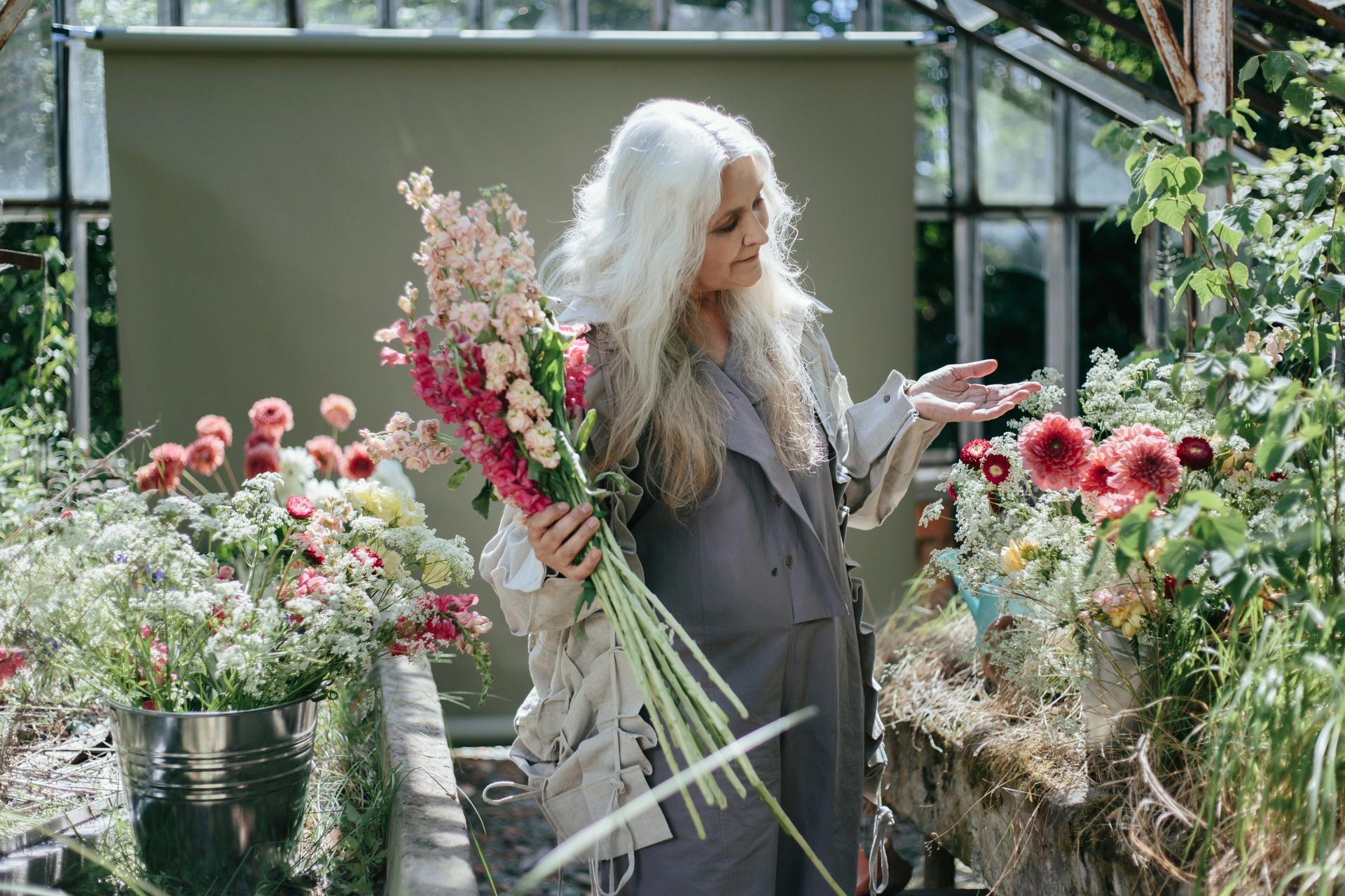 Woman with long white hair, holding flowers in a greenhouse, examining others.