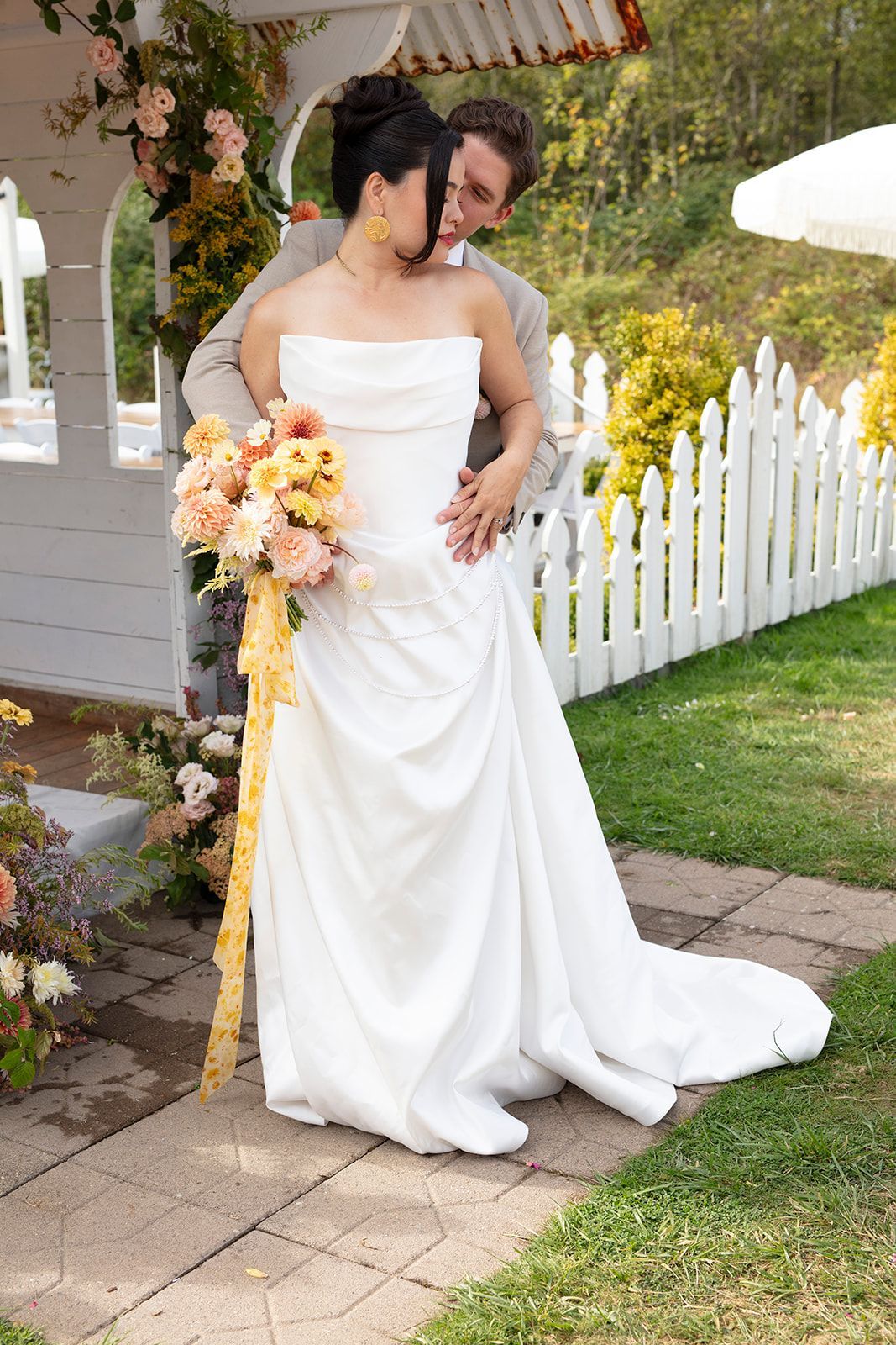 Bride and groom embrace by a white picket fence, woman in white gown holding bouquet, man in suit.
