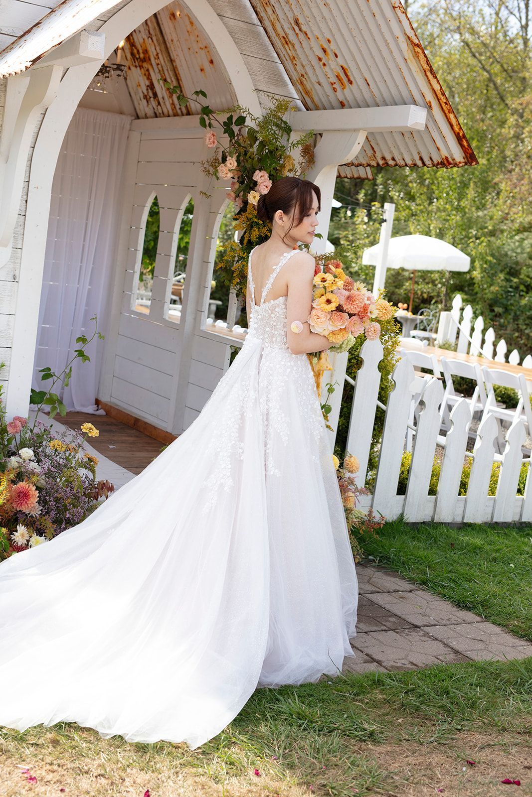 Bride in a white gown stands near a decorative white structure with flowers, holding a bouquet, outdoors.