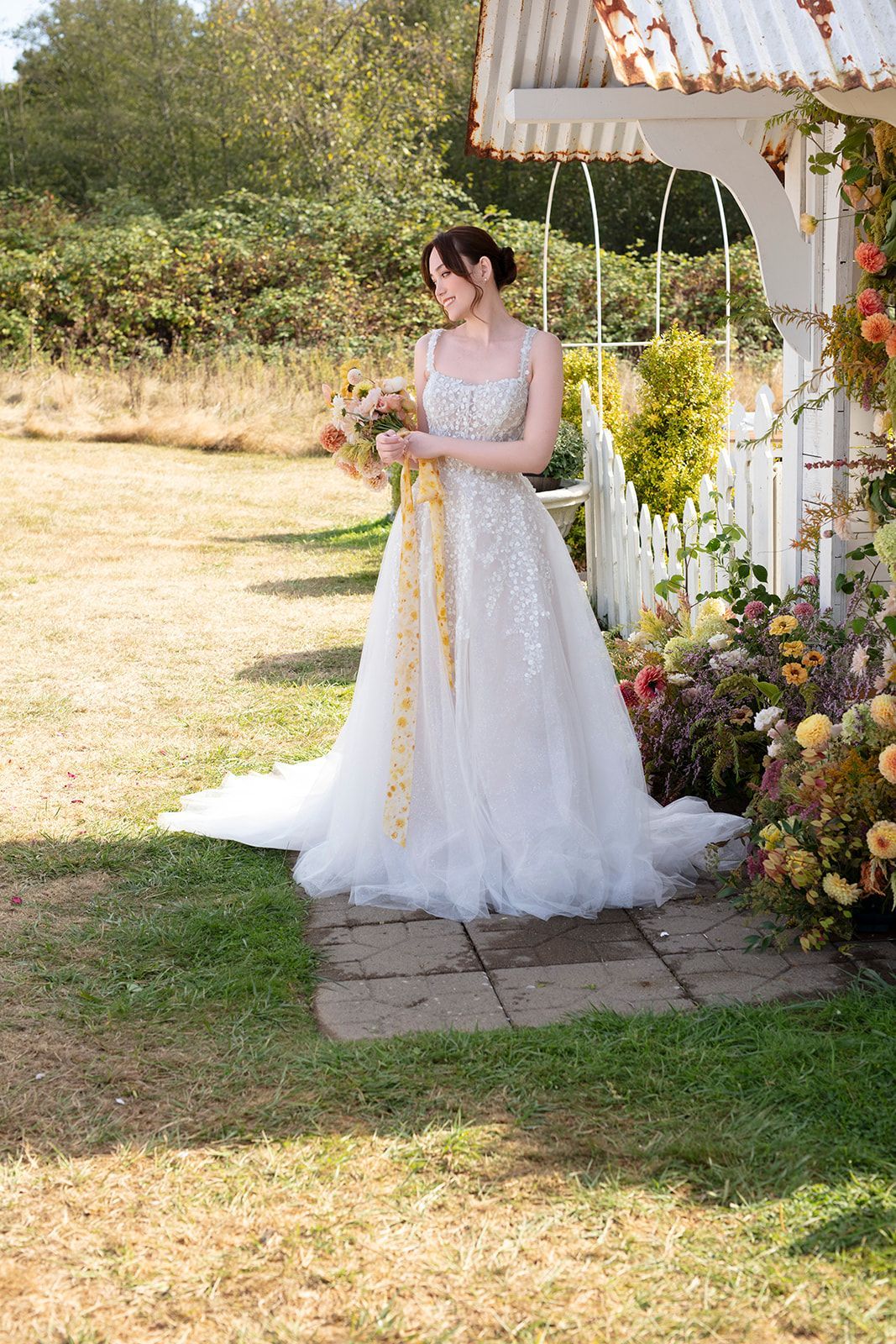 Bride in a sparkly wedding dress holds bouquet with yellow ribbon near a white gazebo in a garden.