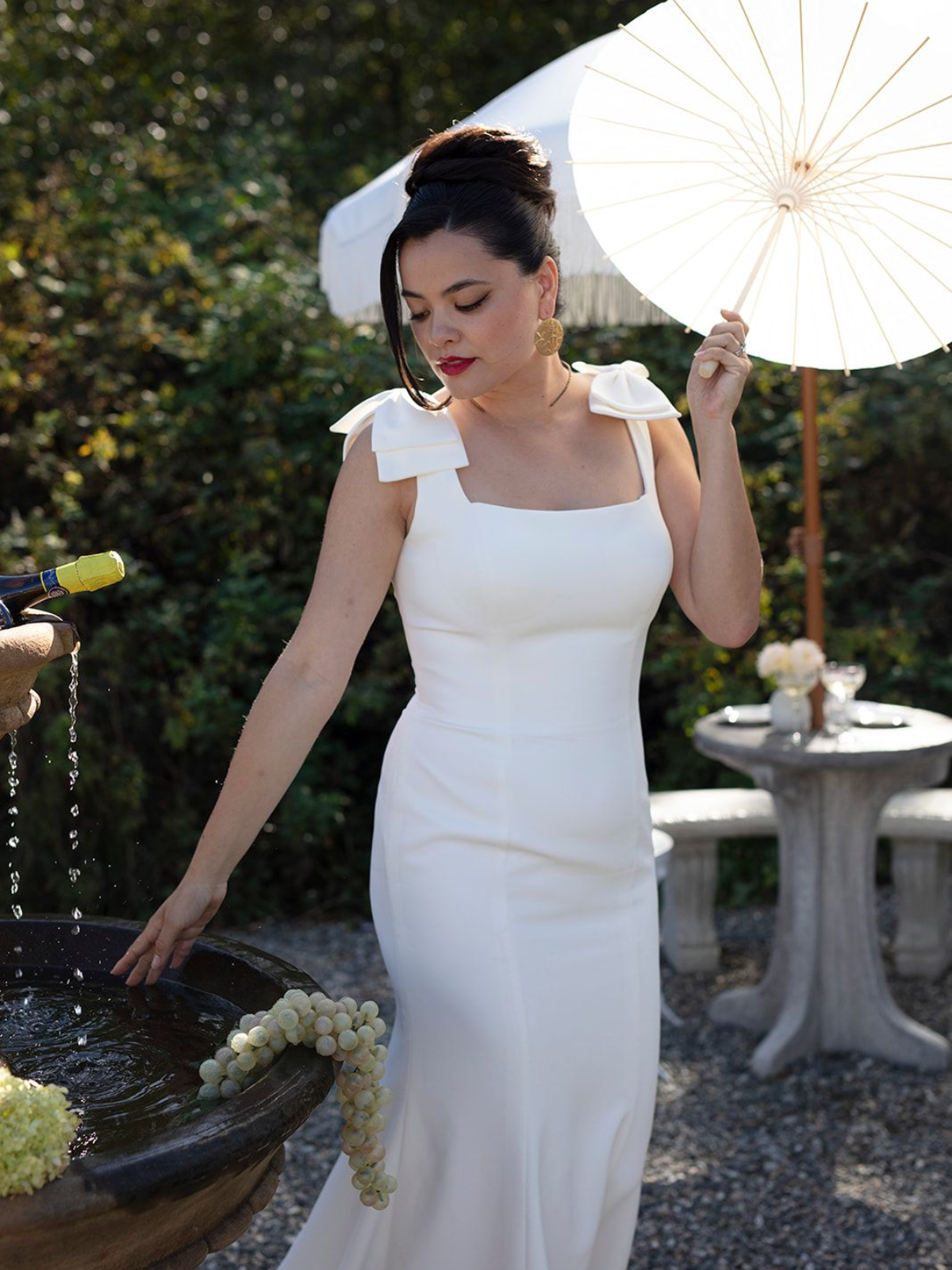 Woman in white dress touches fountain, holding a parasol in an outdoor setting.
