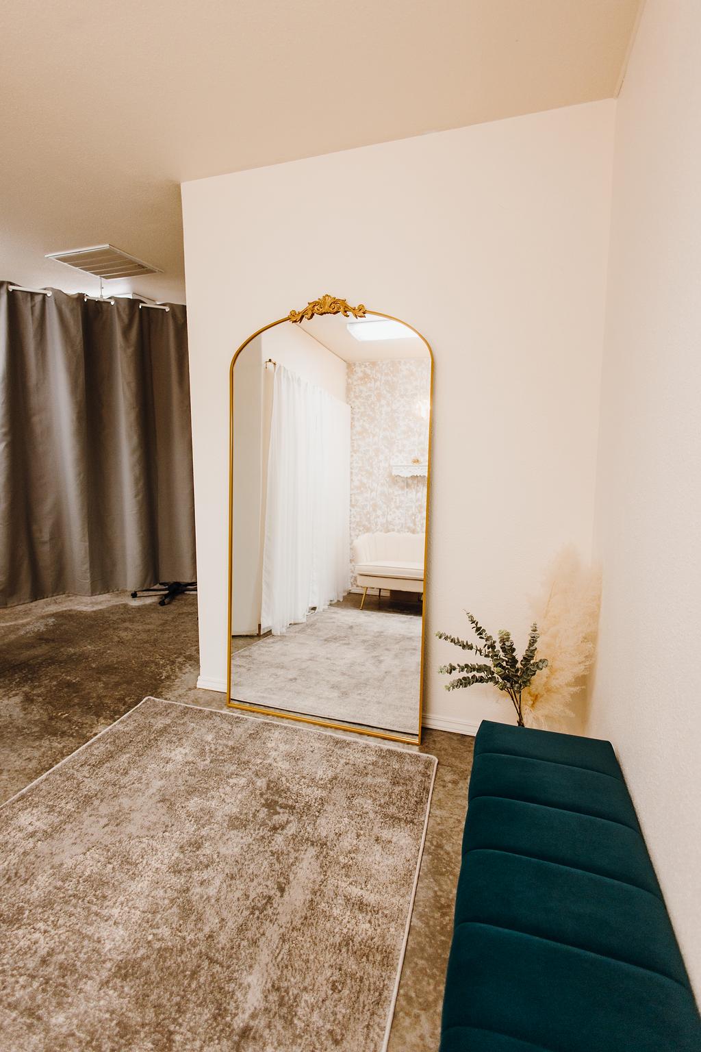 A fitting room with a gold-framed mirror, a teal bench, and a rug. The room has neutral tones.