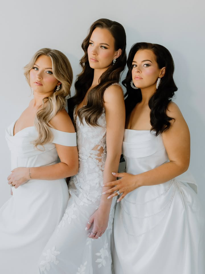 Three women in white gowns pose against a white backdrop; two have dark hair, one blond.