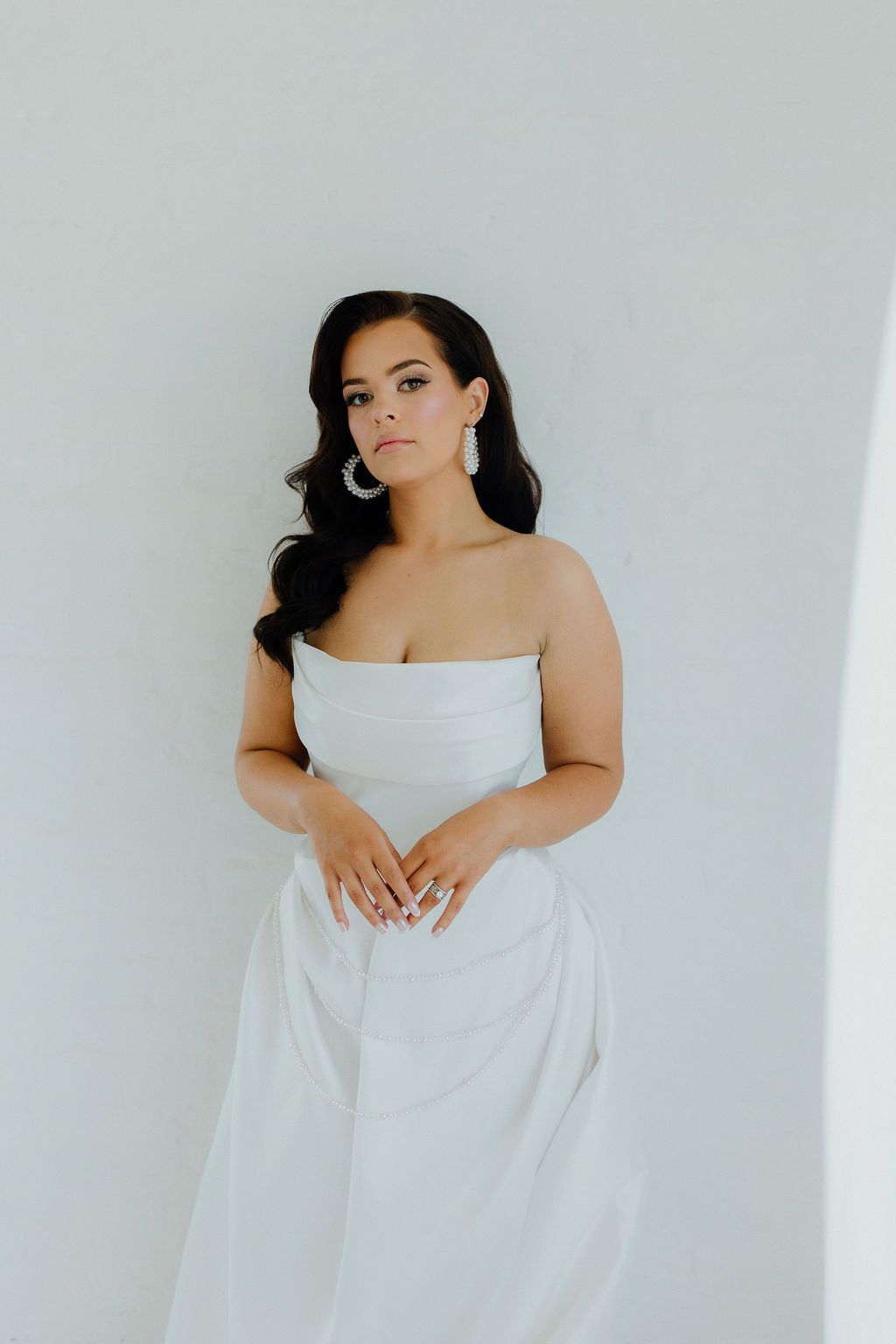 Woman in white strapless dress, hoop earrings, hands clasped, posing against a white wall.