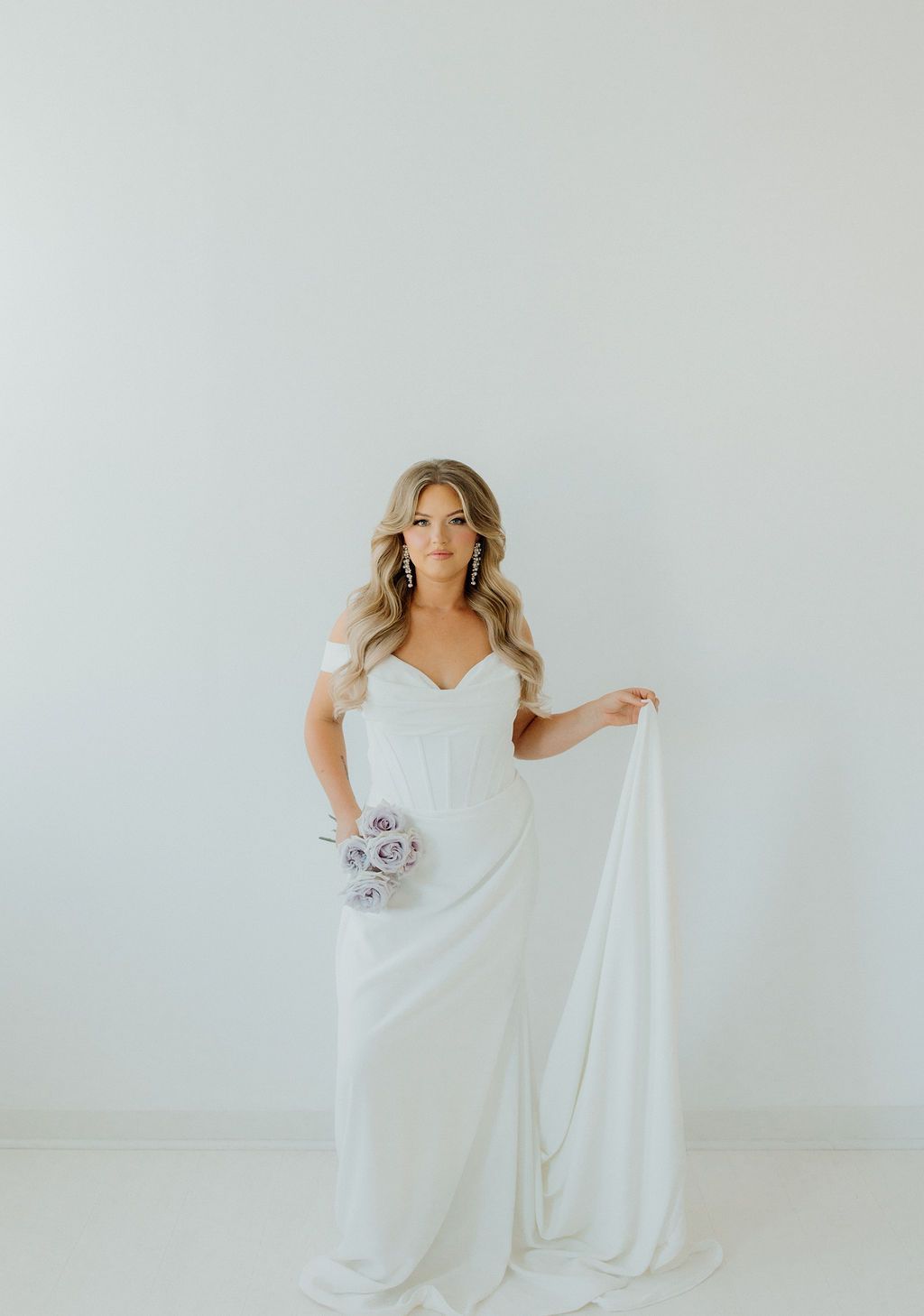 Woman in a white wedding dress holds a bouquet and fabric, standing in front of a white wall.