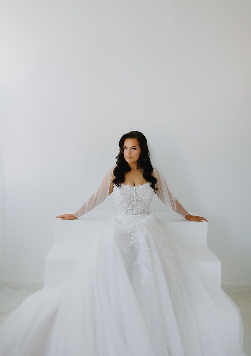 Woman in a white wedding gown sits on white steps, looking towards the viewer.
