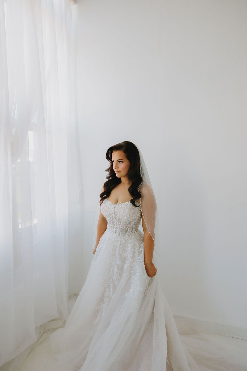 Bride in white wedding dress, holding the skirt, standing in front of a window.