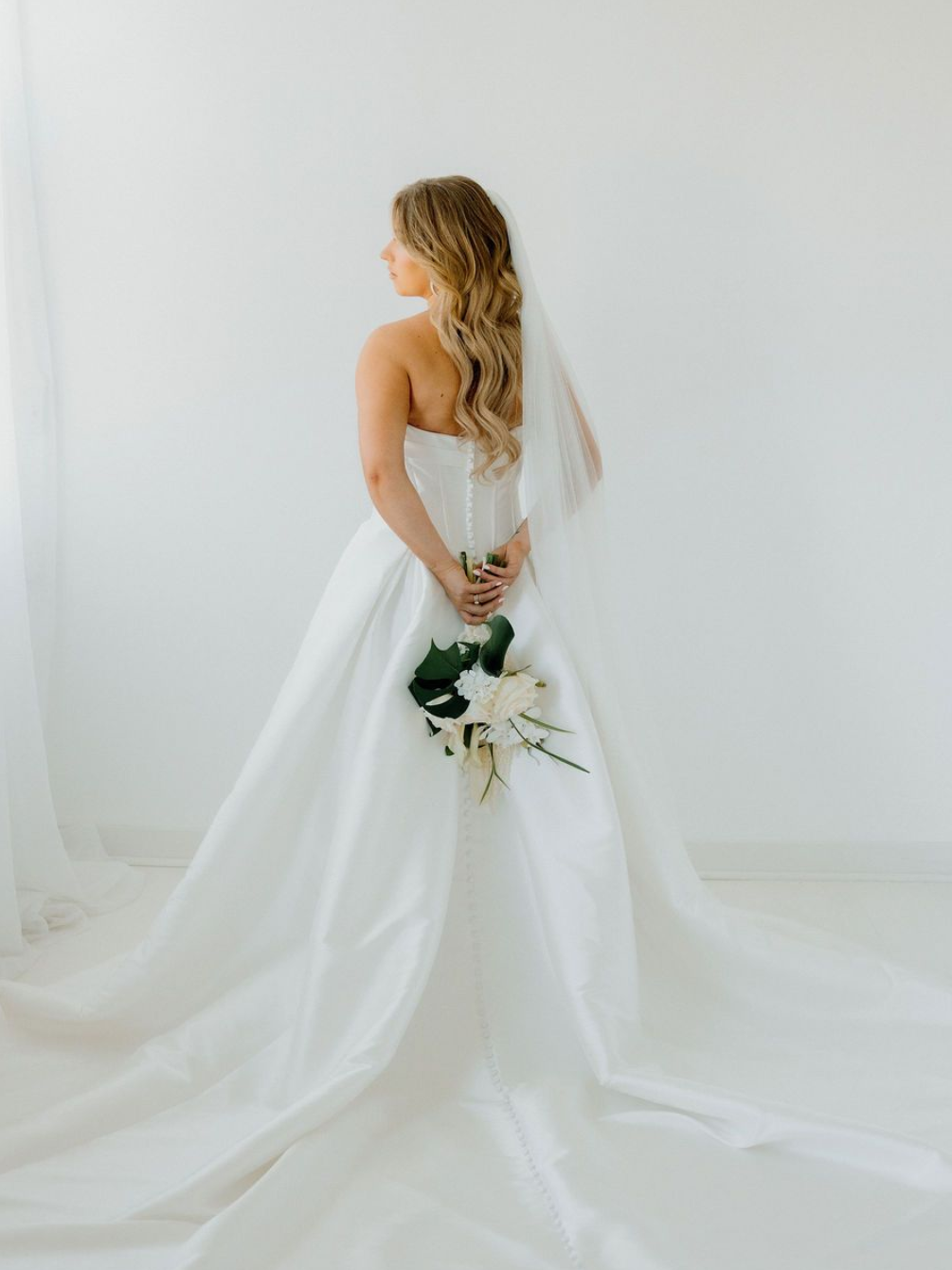 Bride in a white strapless gown, holding a bouquet. She's standing near a white wall, with a veil.