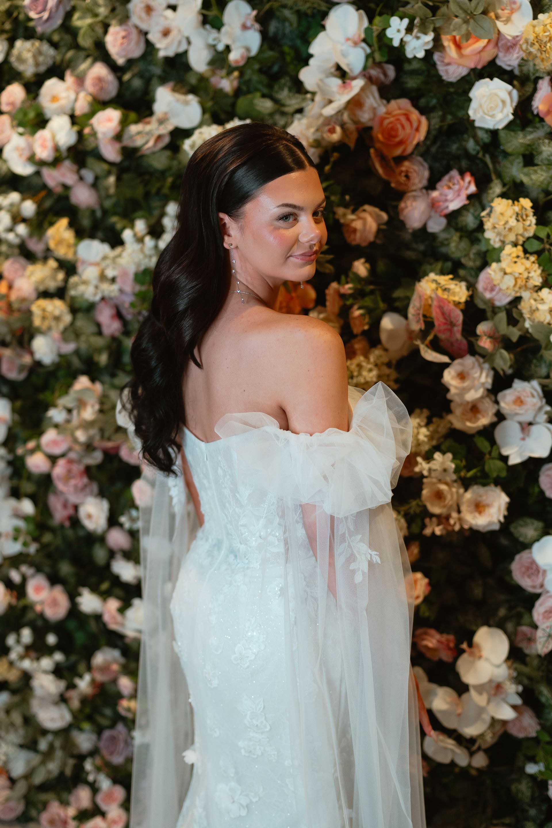 Bride in a white strapless gown, holding a bouquet. She's standing near a white wall, with a veil.