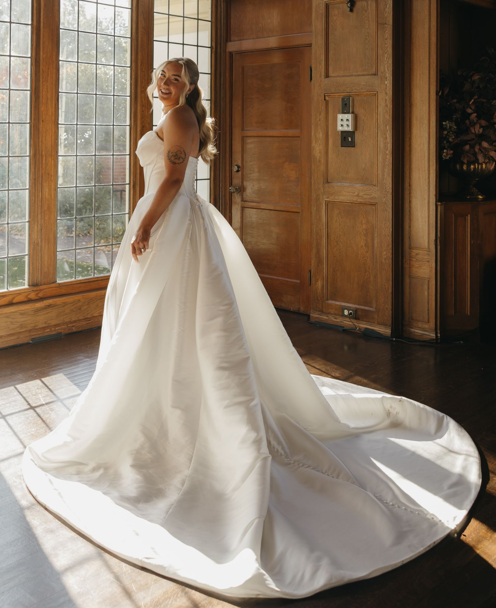 Woman in wedding dress stands near large window in a wood-paneled room.