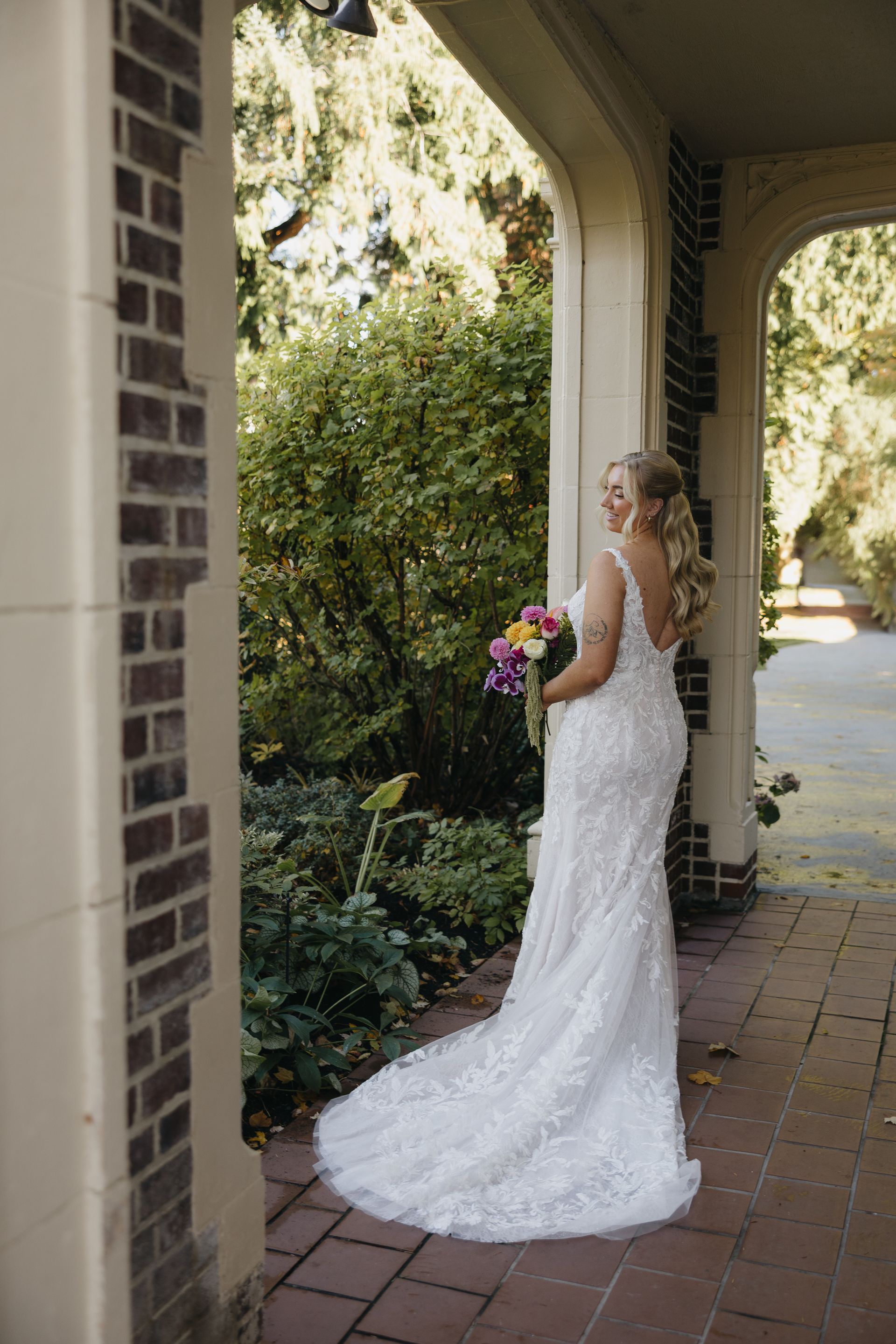 Bride in a white, floral lace gown, holding a bouquet. Standing under a brick and stone archway.