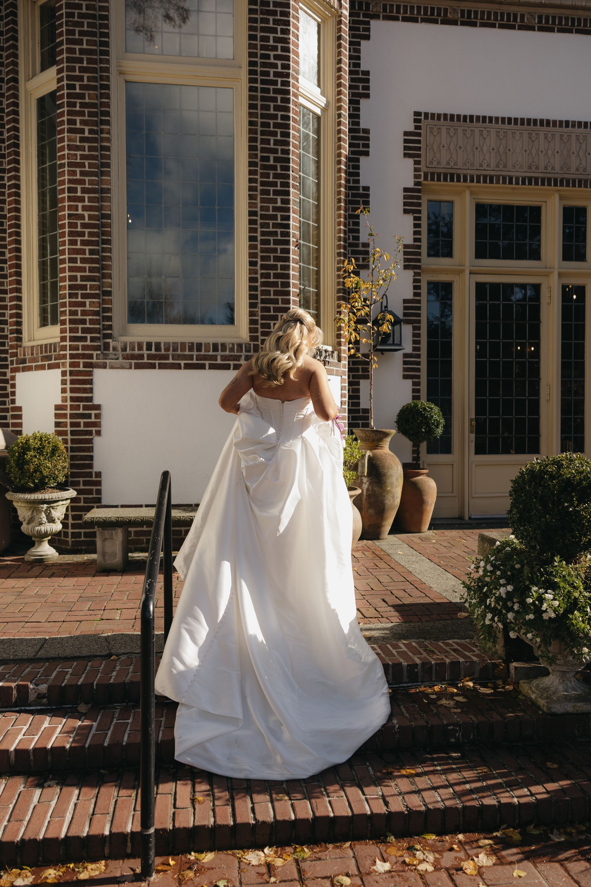 Bride in white wedding dress walks up brick steps towards a building with large windows.