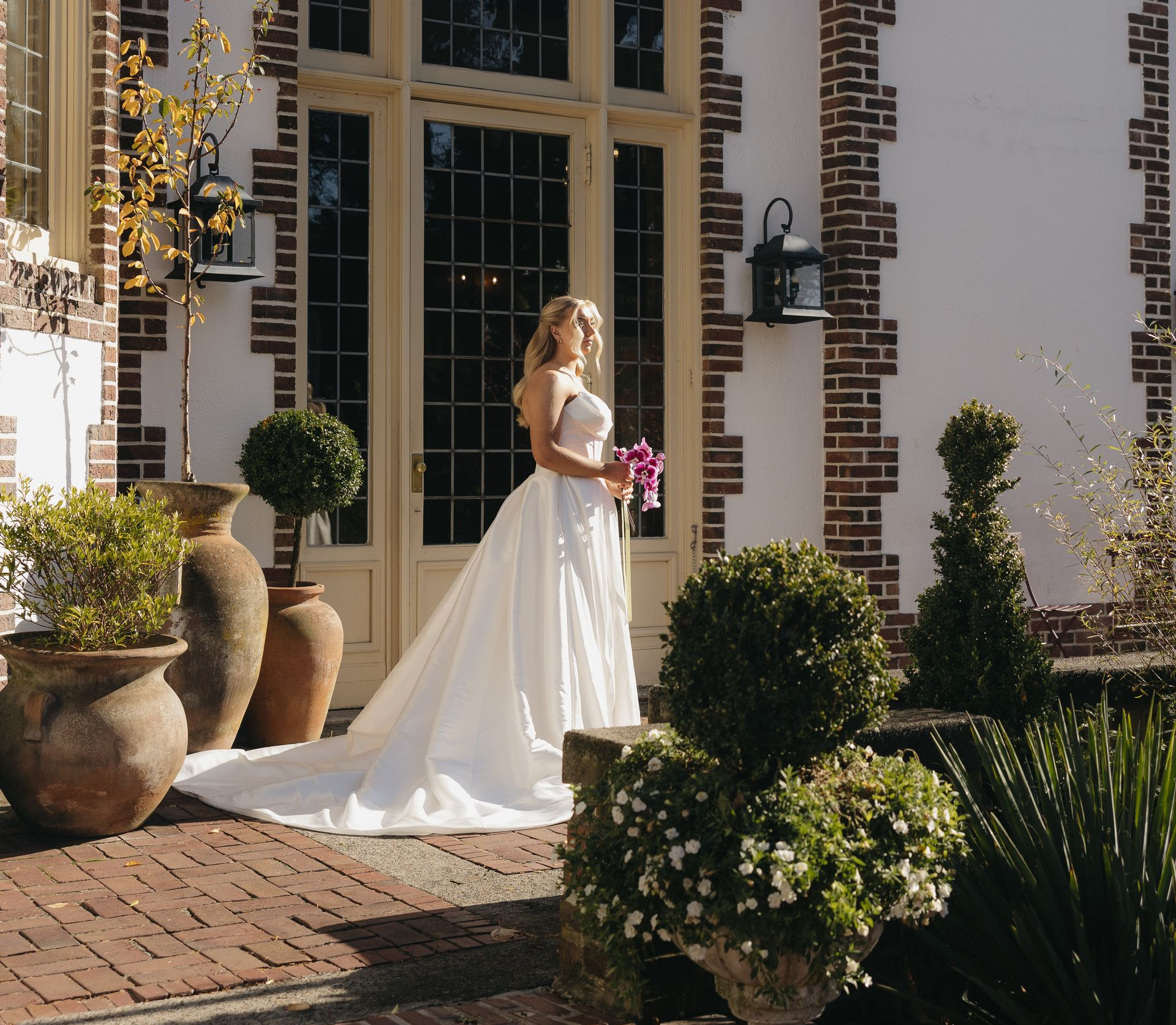 Bride in a white gown holding flowers, standing near a building with a large window and potted plants.