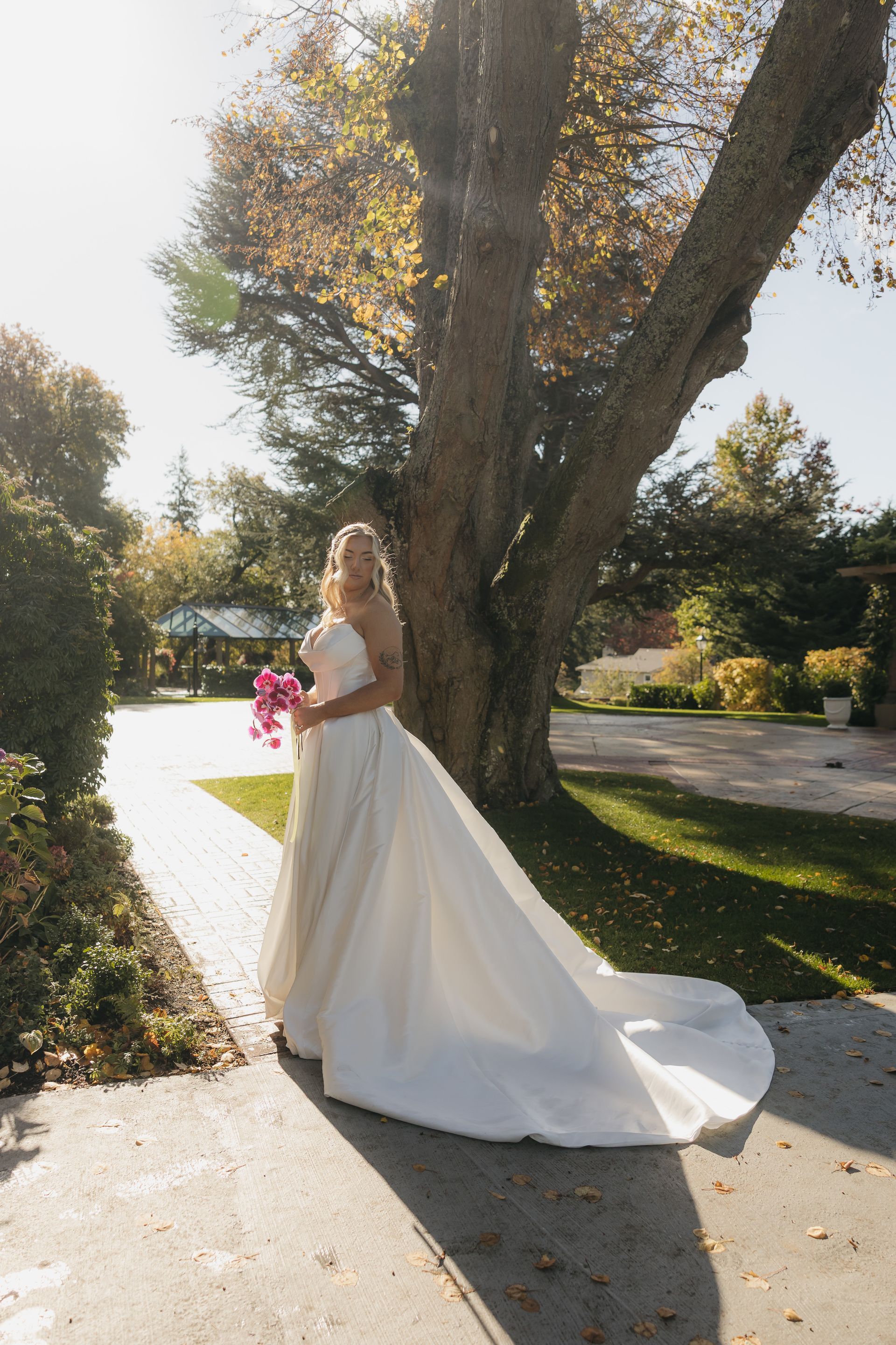 Bride in a white gown stands holding a pink bouquet near a large tree in a sunny outdoor setting.