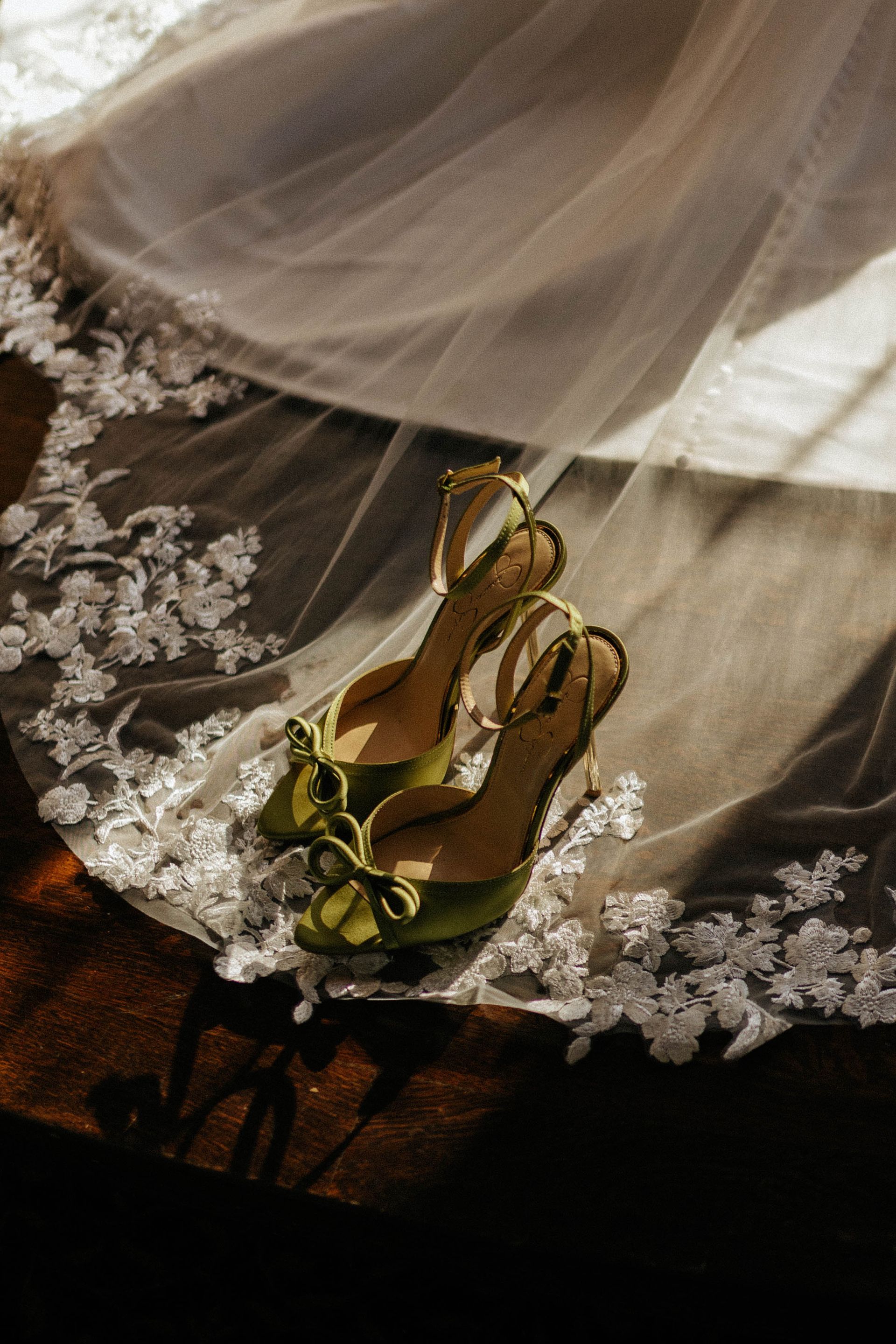 Green high-heeled shoes with bow detail sit under a white lace veil on a wooden surface.