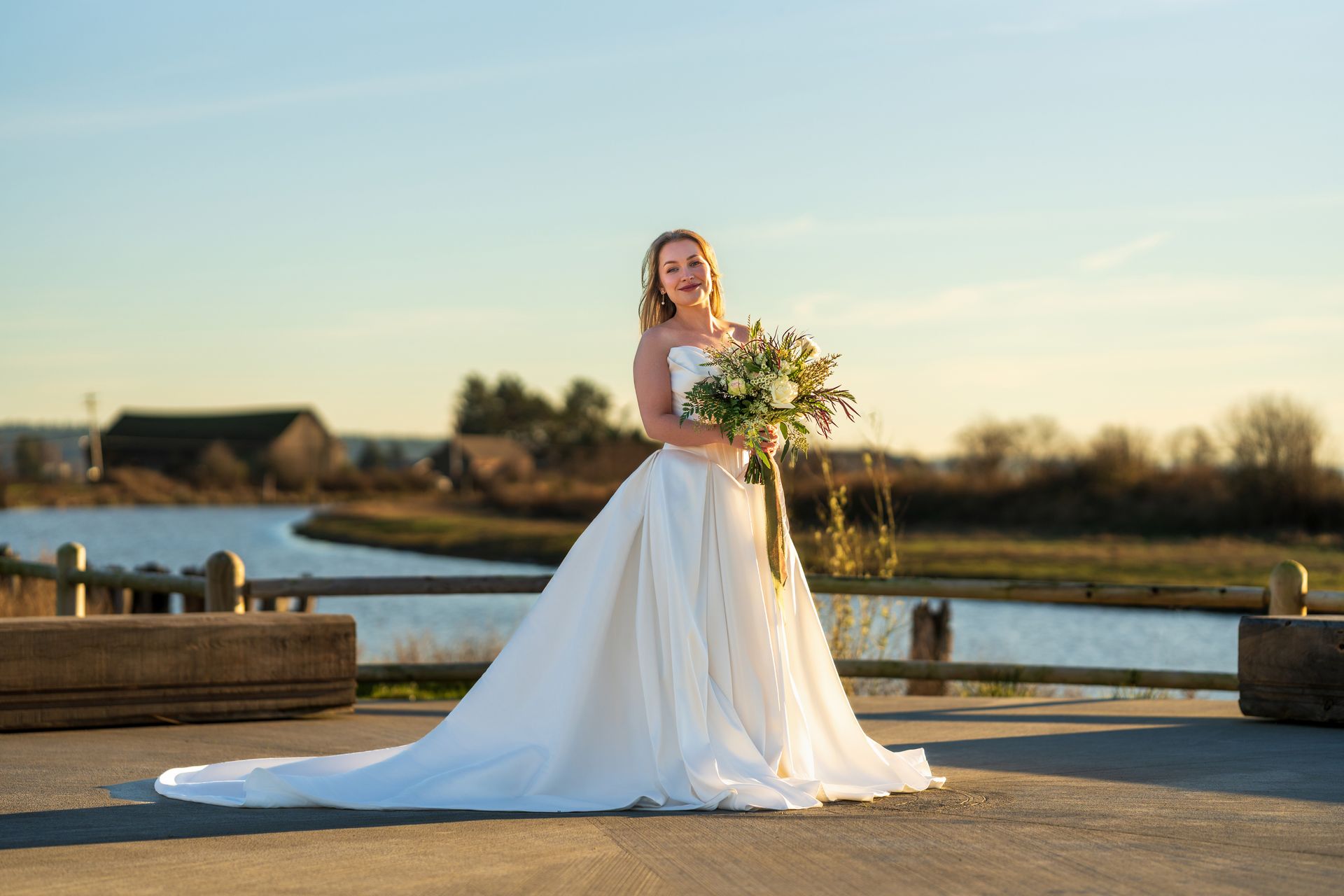 Bride in a white gown holding flowers, standing near a building with a large window and potted plants.