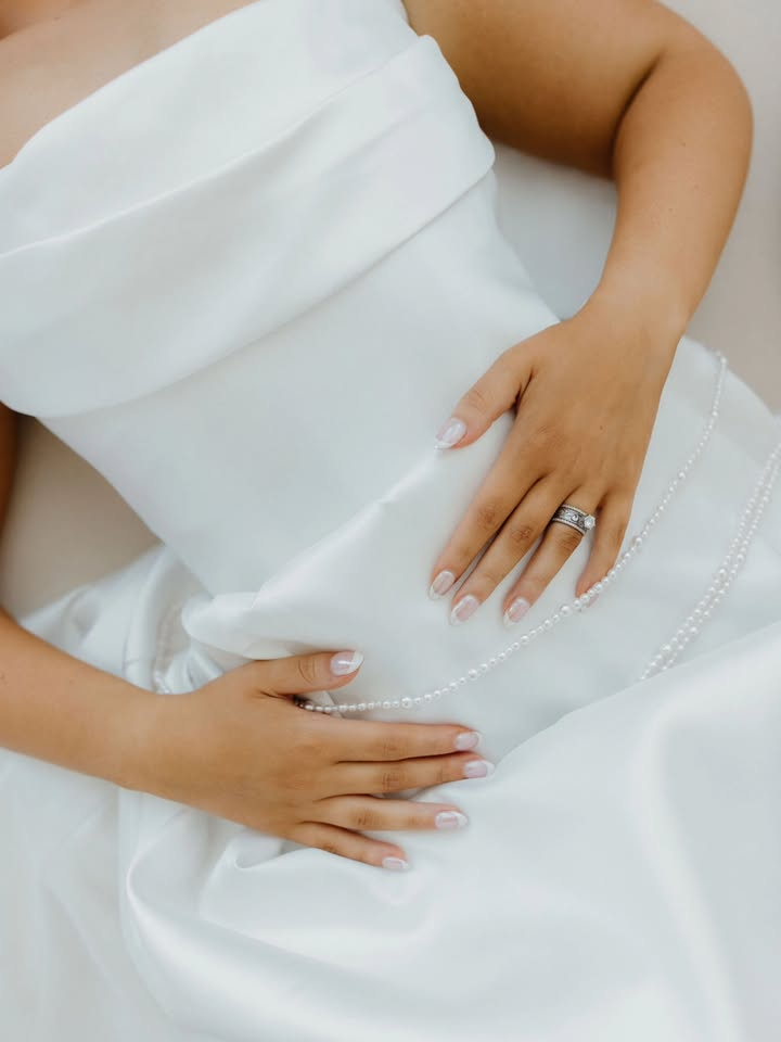 Bride wearing a white wedding dress, hands resting on the stomach, showing off a ring.
