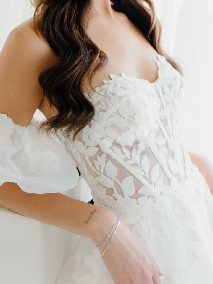 Woman in a white floral wedding dress with off-the-shoulder sleeves, leaning against a white surface.