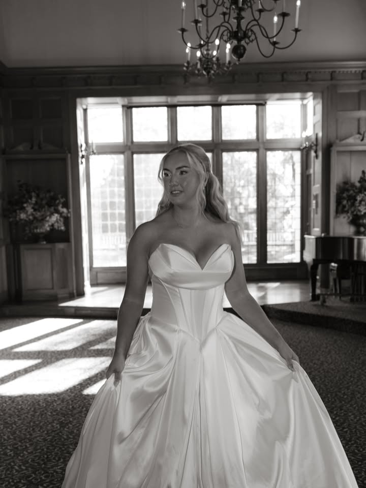 Woman in a strapless gown in a ballroom with large windows and a chandelier.