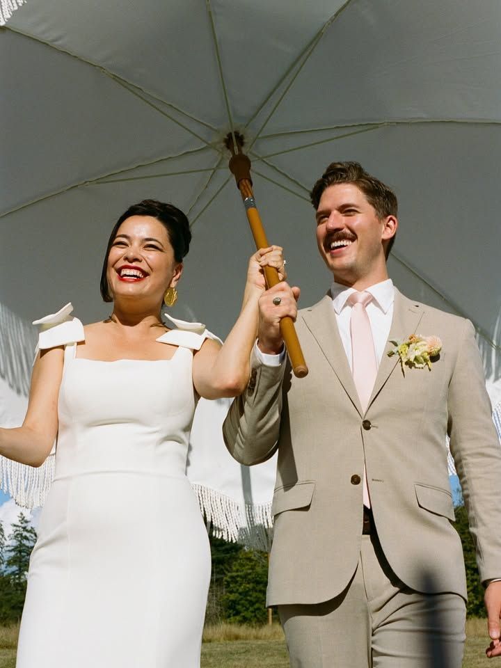 Couple smiles, holding umbrella. Woman in white dress, man in tan suit, outdoors.