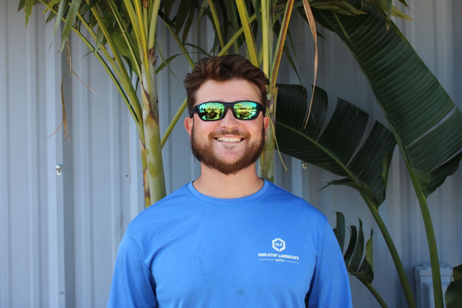 Man in sunglasses smiles, wearing a blue shirt, in front of green plants and a white wall.