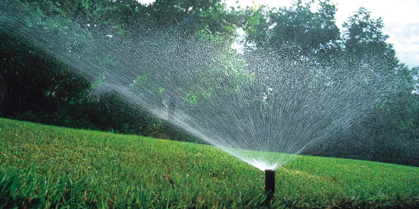 A sprinkler watering green grass in a yard, with a backdrop of trees and a cloudy sky.
