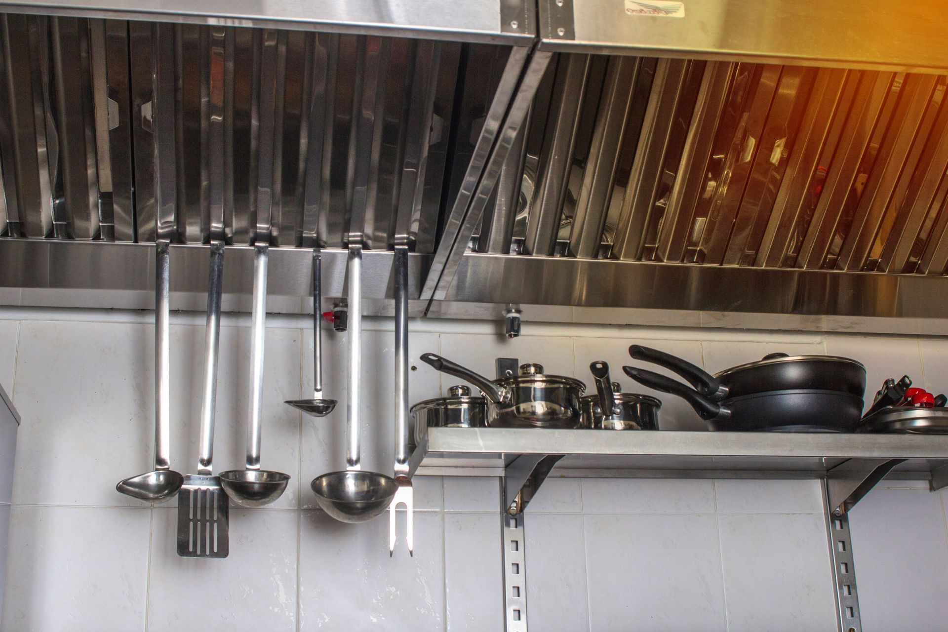Kitchen vent hood above a shelf with hanging ladles, pots, and pans against a tiled wall.