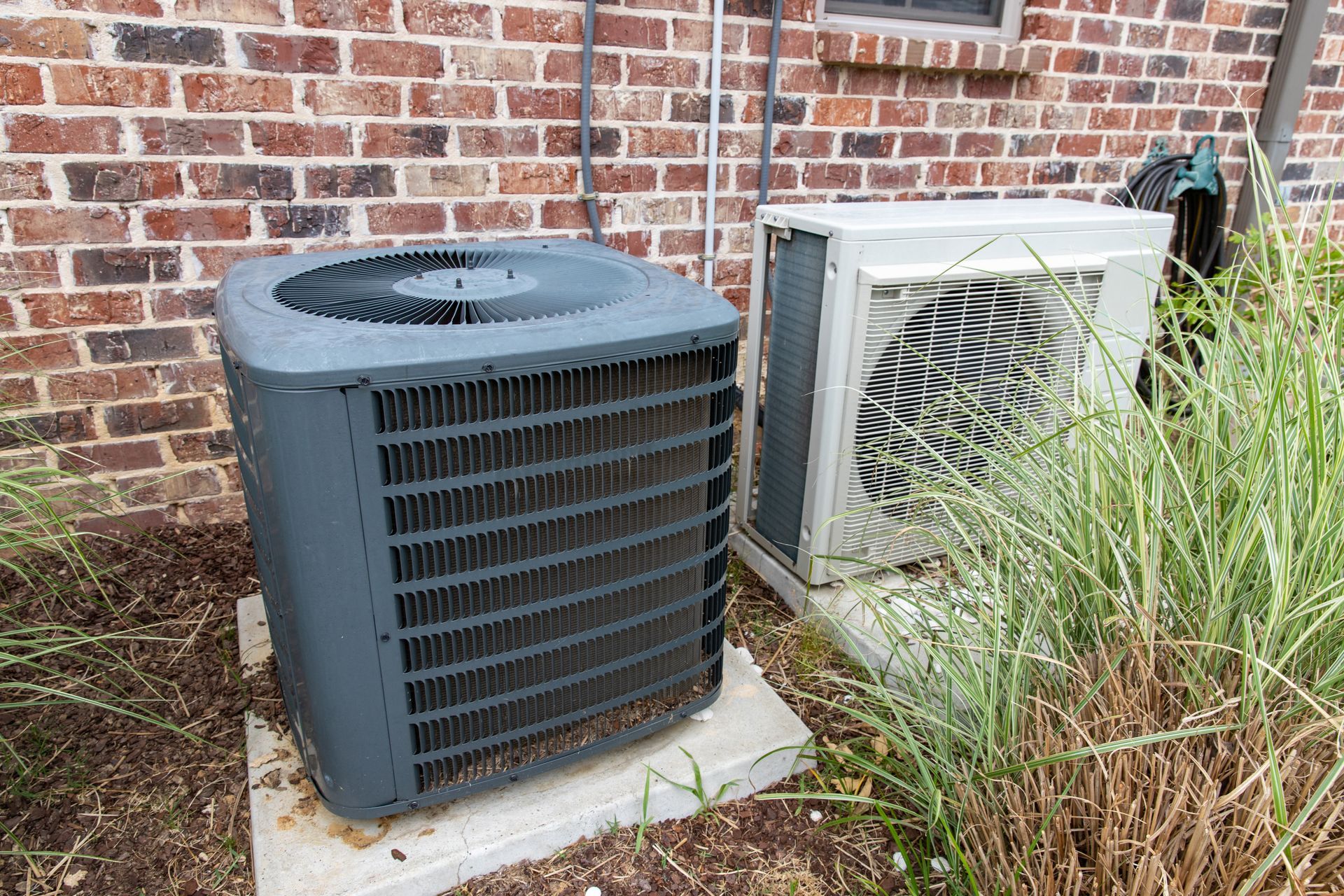 Two outdoor air conditioning units on concrete pads next to a brick wall and tall grass.