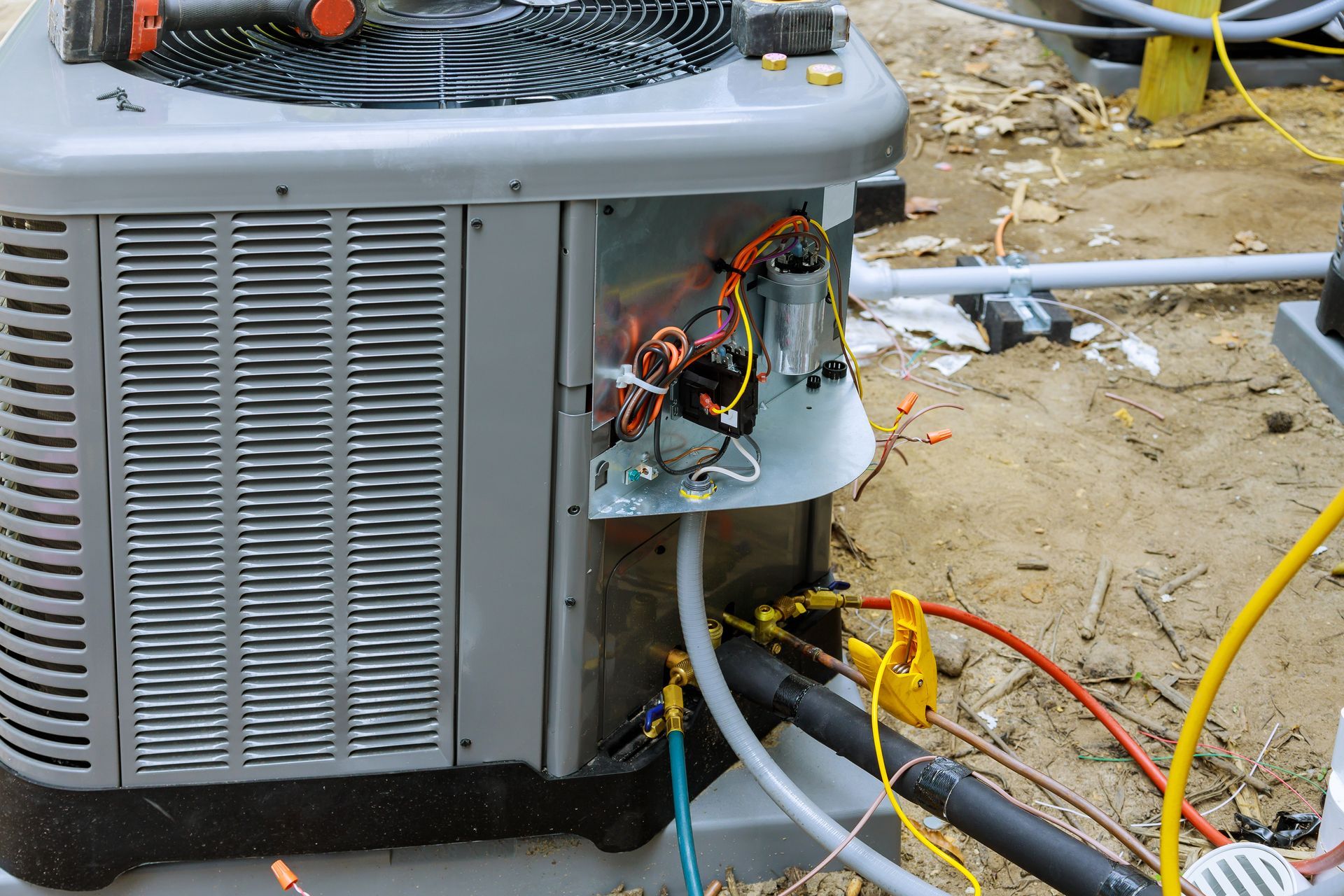 A gray outdoor air conditioning unit with its electrical panel open and service hoses connected for maintenance.