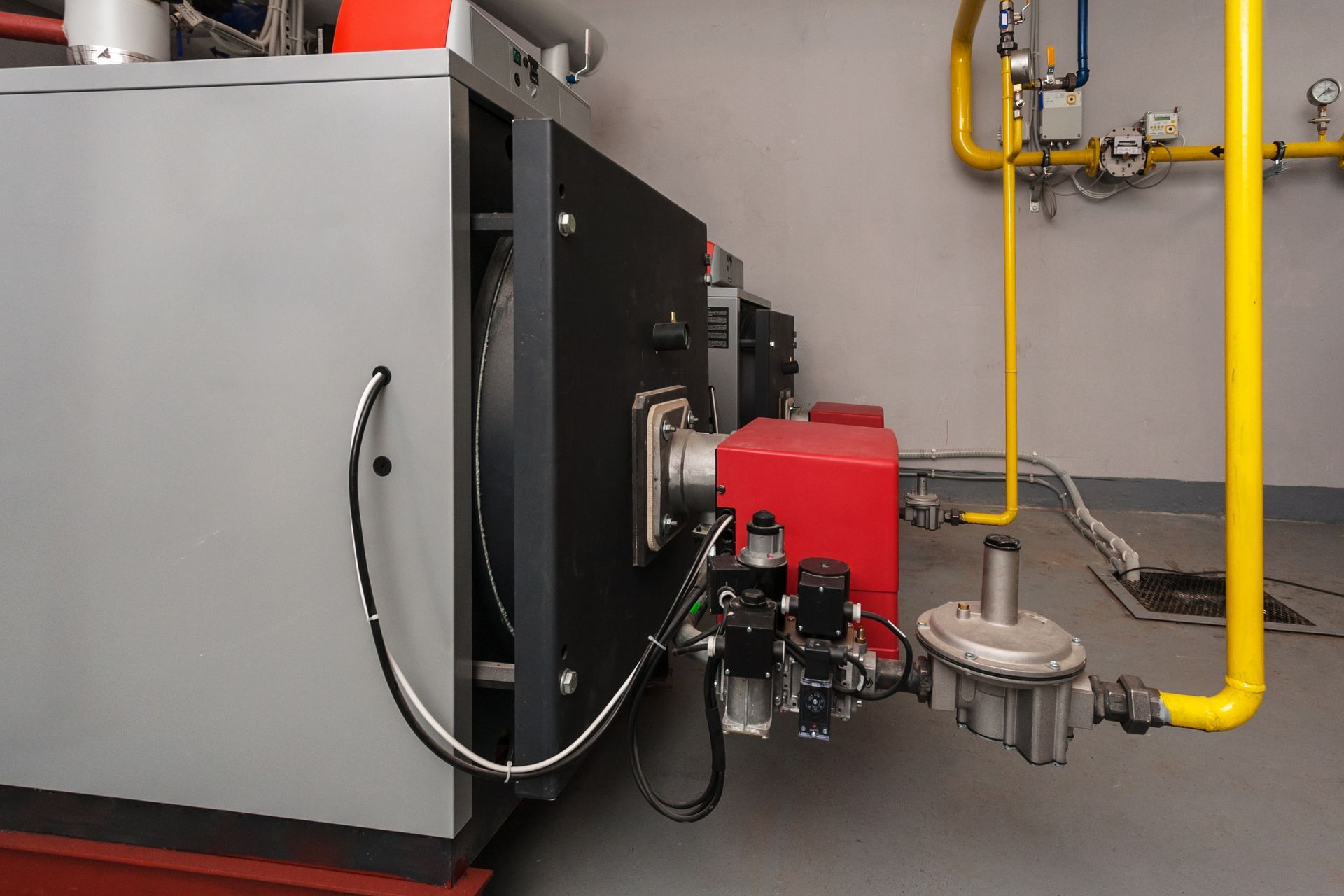 A large industrial boiler with a red burner unit connected to yellow gas supply pipes in a utility room.