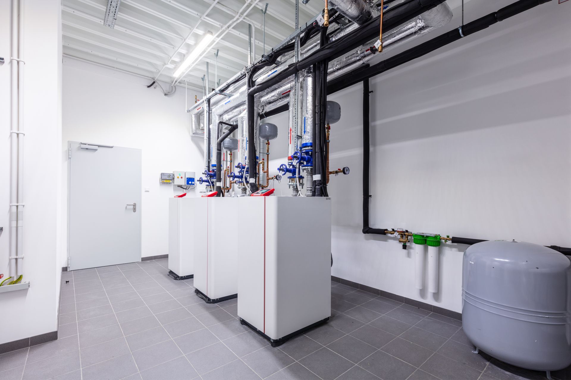 A mechanical room with three white industrial heating units, overhead black pipes, and a grey pressure tank on tiled floors.