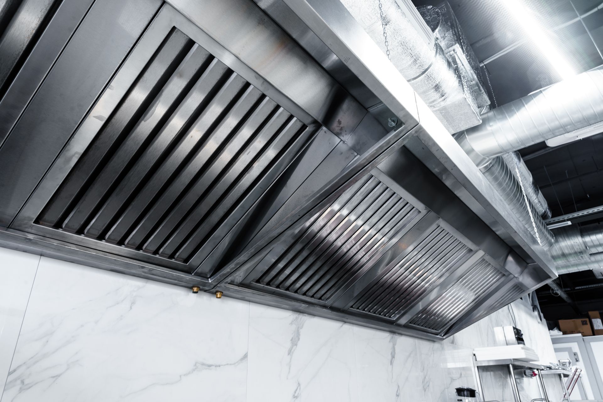 A wide, stainless steel commercial kitchen exhaust hood with grease baffle filters mounted above a marble-tiled wall.
