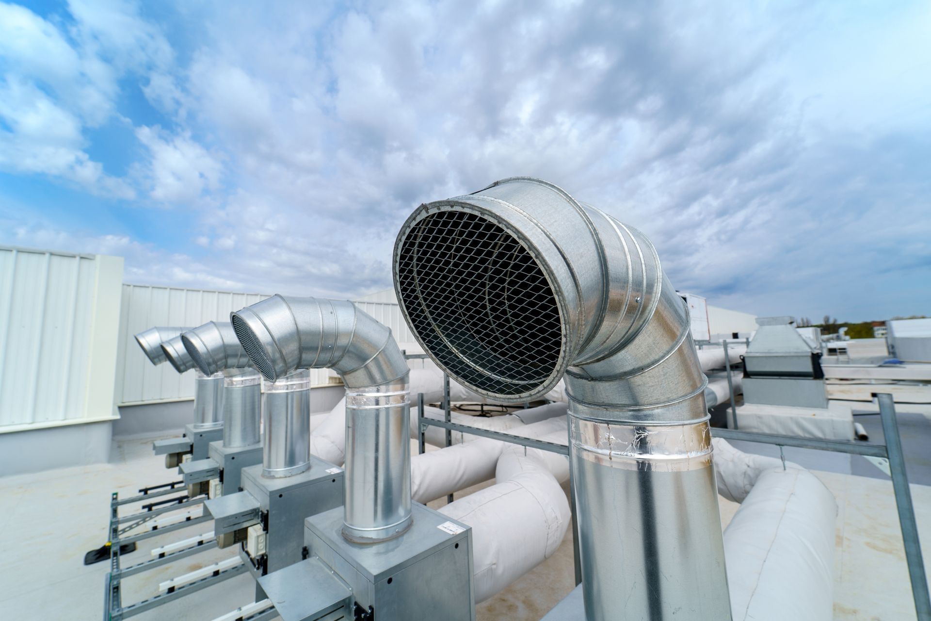 A row of silver, industrial exhaust vents on a flat rooftop against a cloudy blue sky.