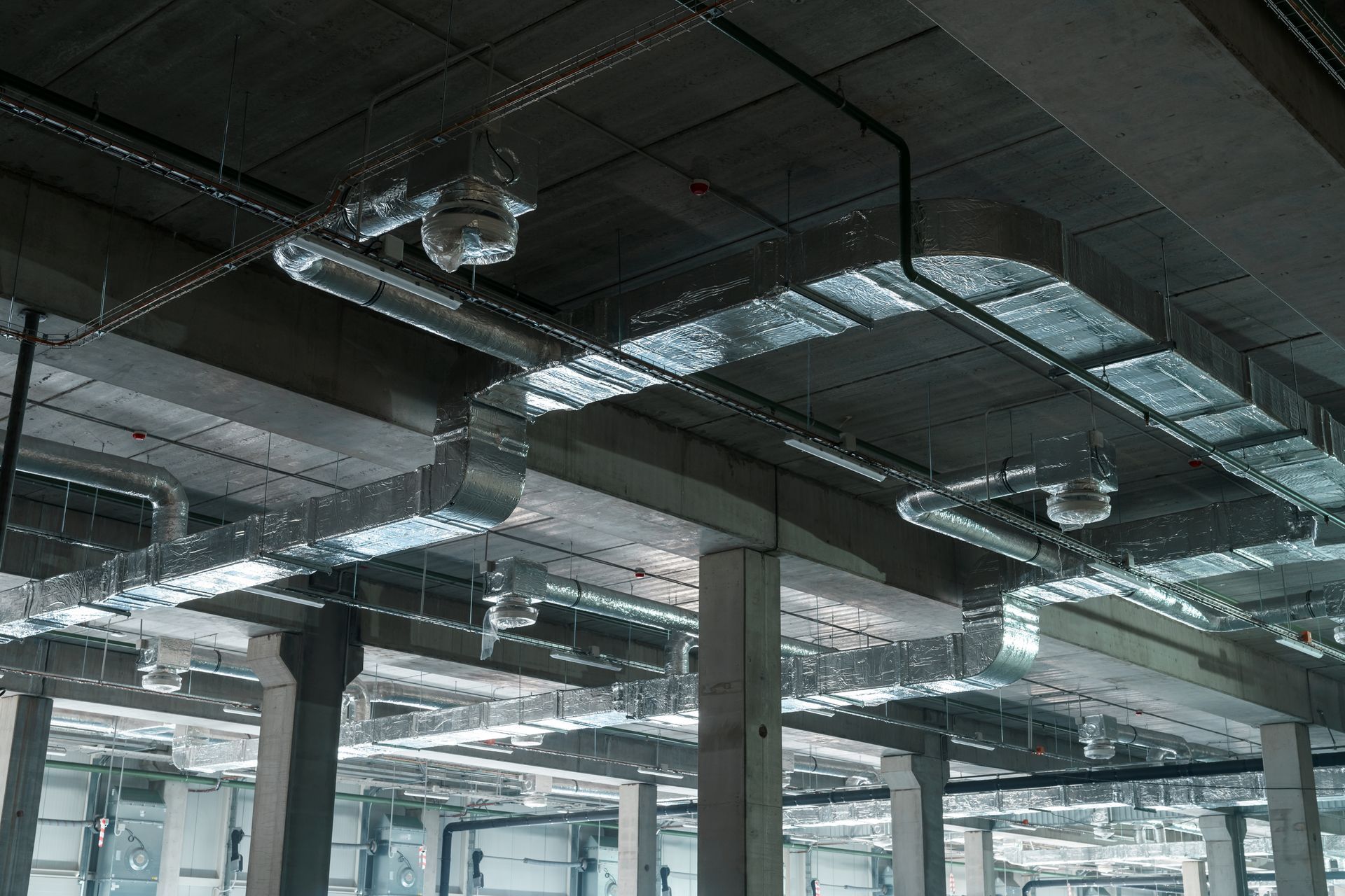 Industrial ceiling with exposed silver HVAC ductwork suspended below concrete beams in an unfinished building.