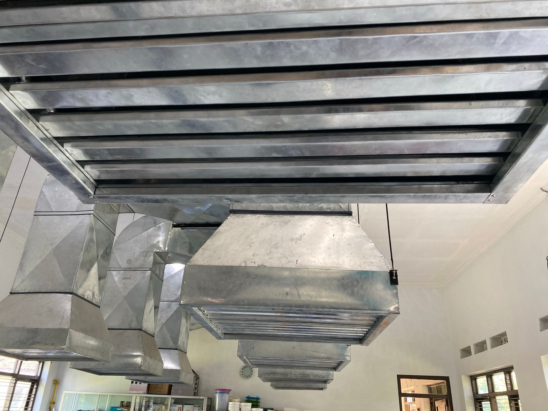 A low-angle view of industrial metal ventilation hoods and ductwork hanging from a kitchen ceiling.