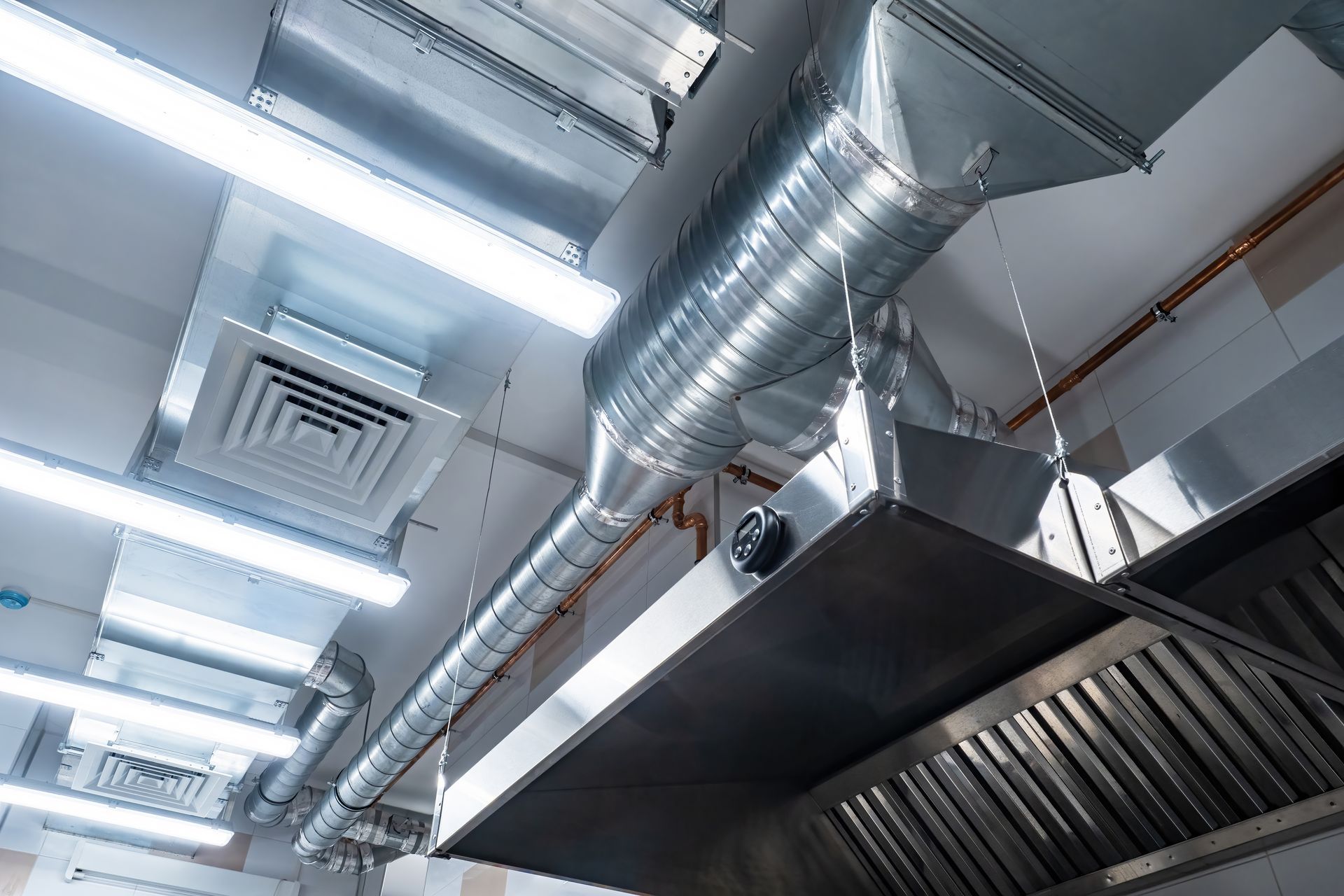 Metal ventilation ducts and a large commercial exhaust hood hanging from a ceiling with bright fluorescent lights.