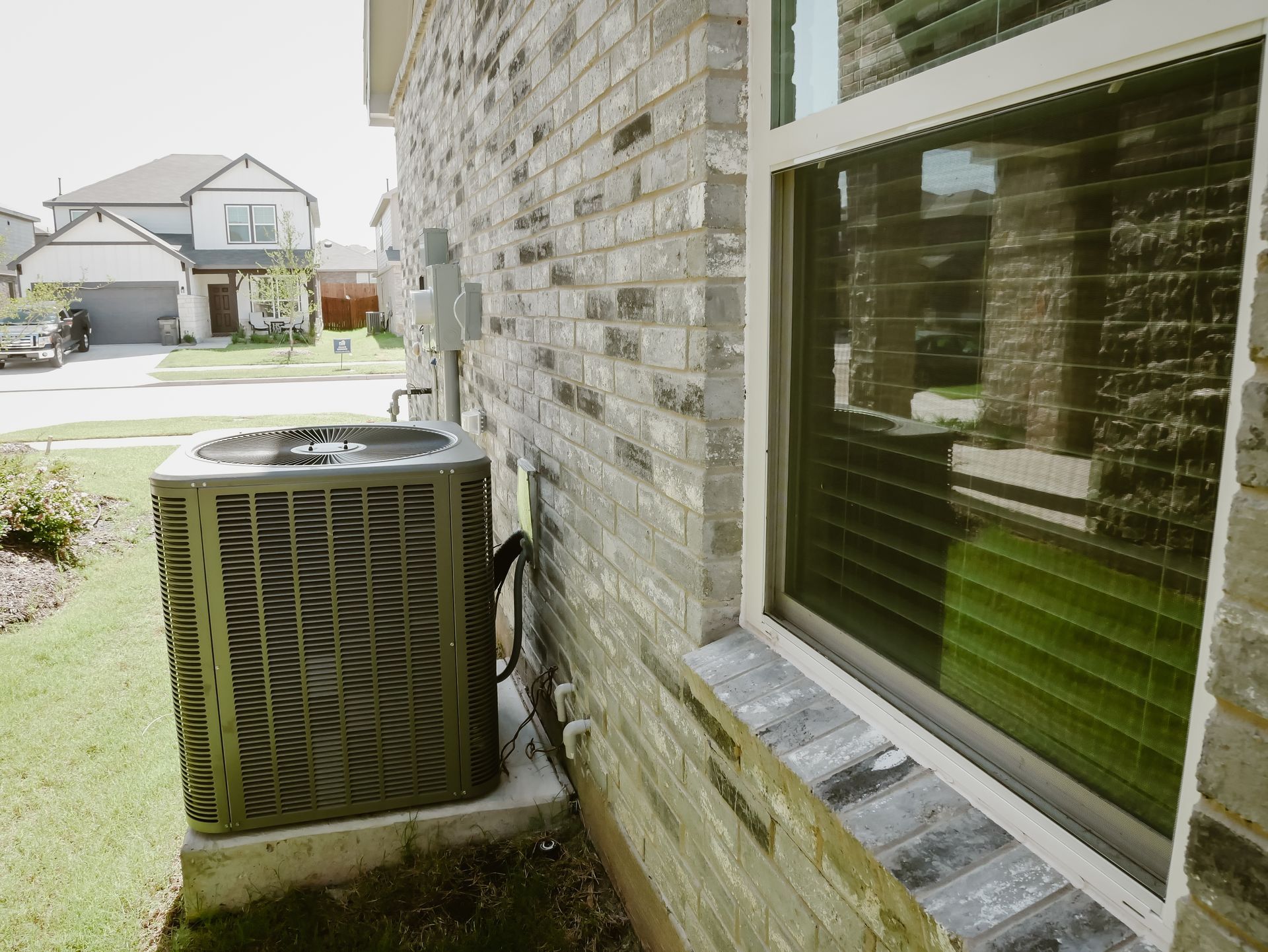 Air conditioning unit next to a brick wall with a window; green grass and a house in the background.