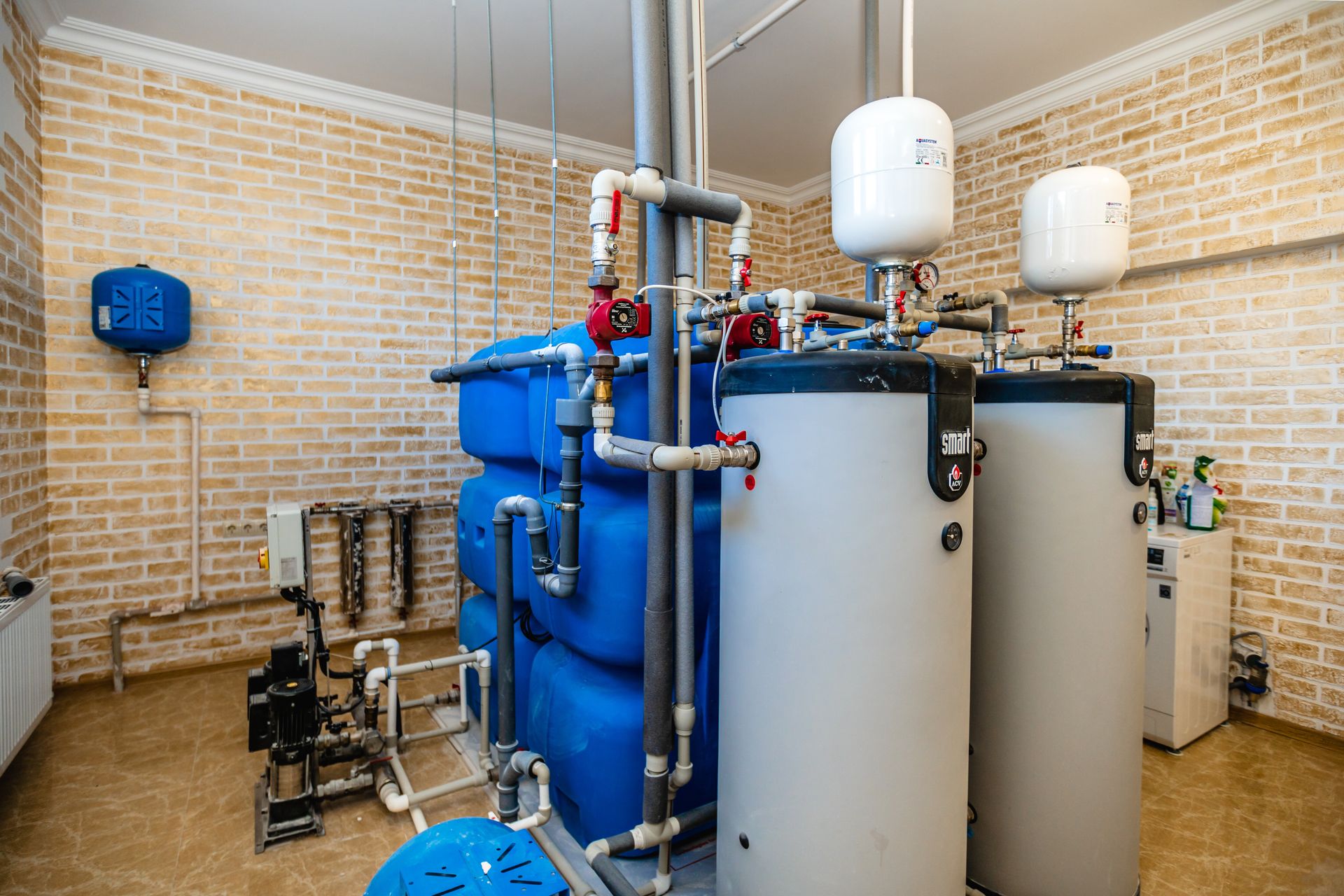 A boiler room with two gray water heaters, large blue storage tanks, and various pipes against a light brick wall.