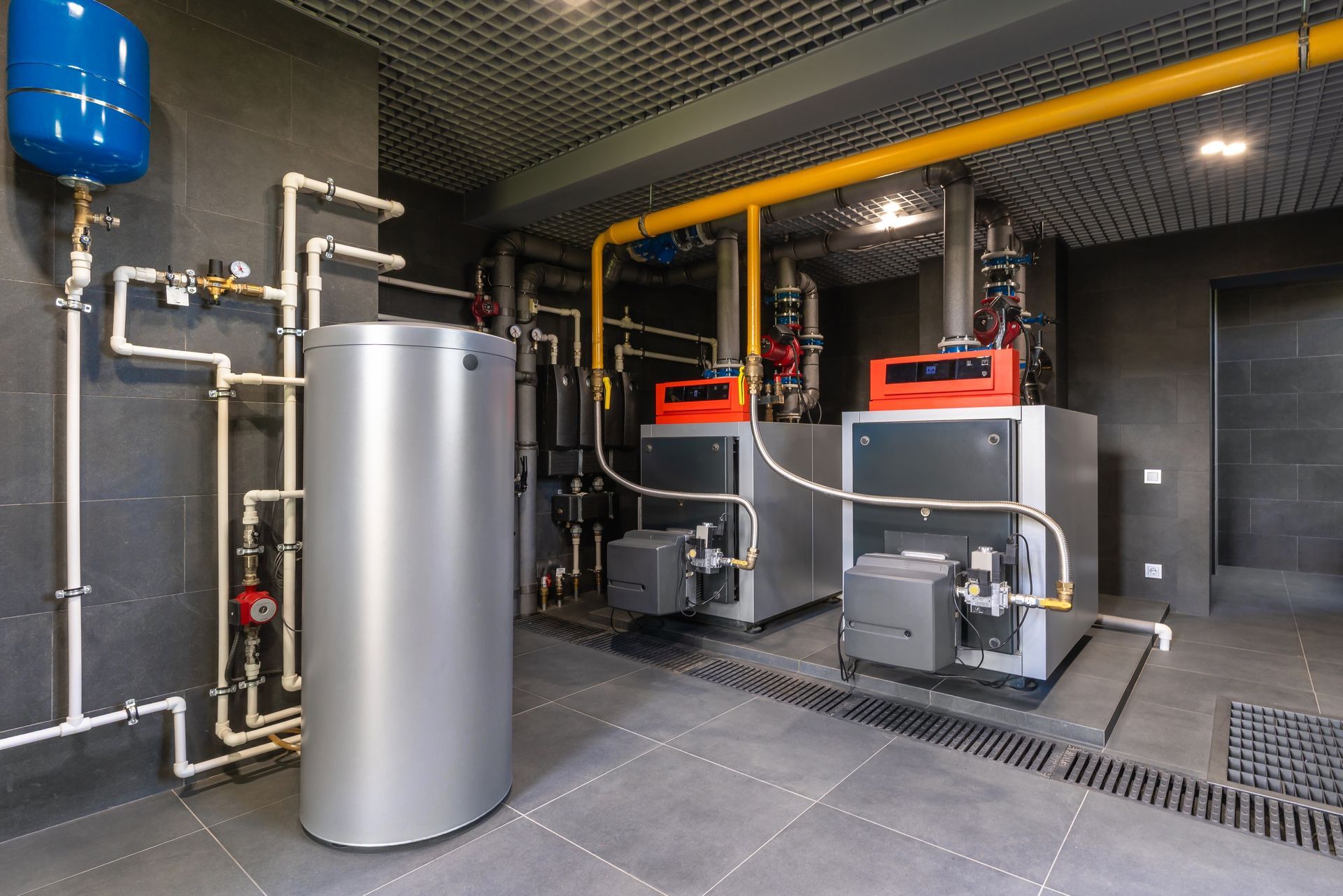 Two industrial boilers and a large cylindrical water tank in a modern mechanical room with dark tiled walls and floors.