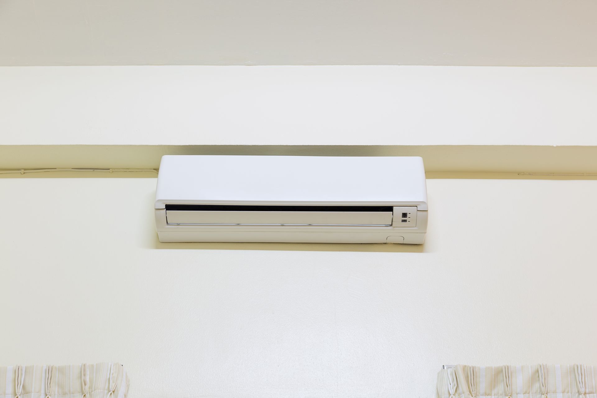 Wall-mounted white air conditioner on a beige wall above patterned curtains