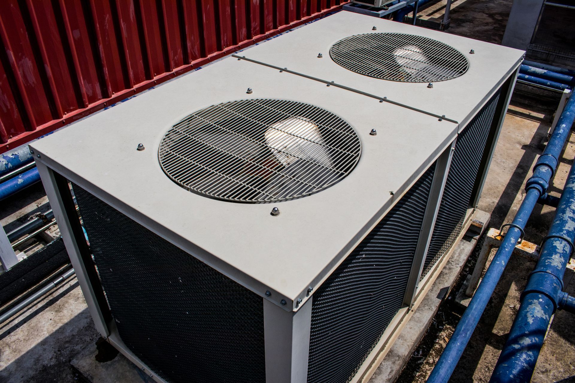An outdoor industrial HVAC unit with two large fans sits on a roof next to blue pipes and a red wall.
