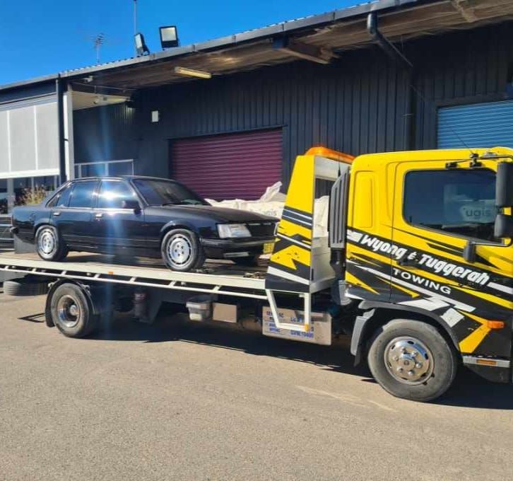 A Yellow Towing Truck is Carrying a Black Car — Wyong and Tuggerah Towing Service in Tuggerah, NSW