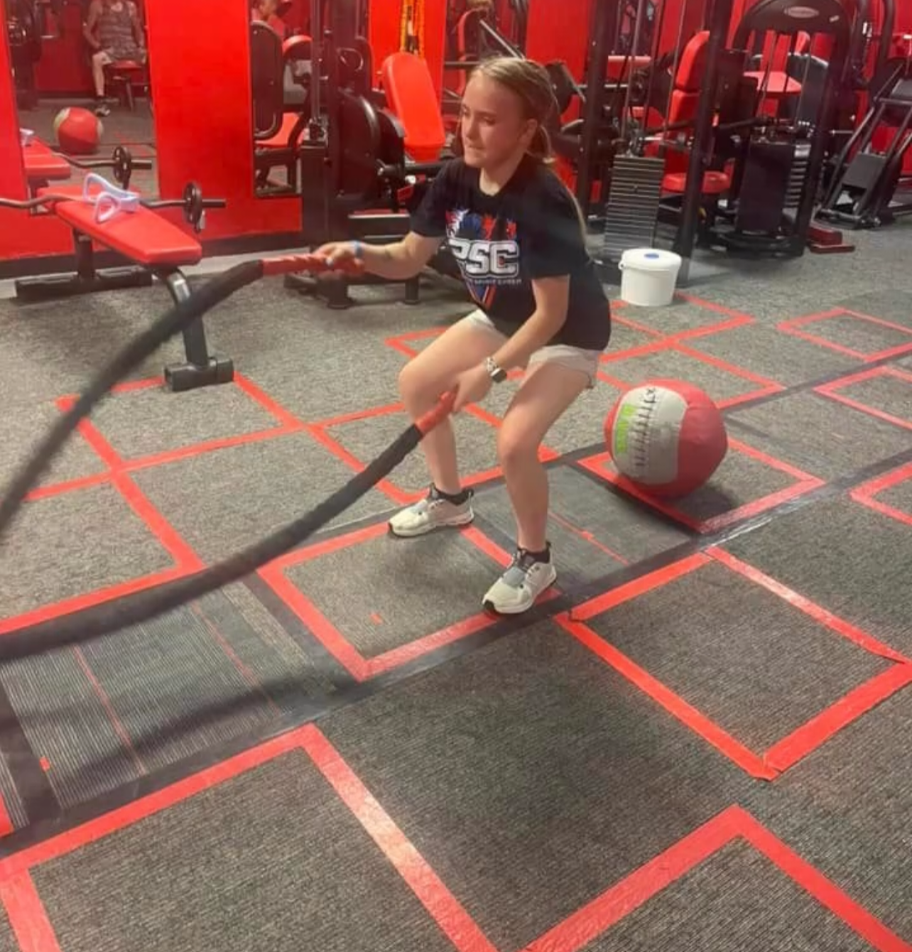 Young woman in a gym using battle ropes, performing a squat. Red and gray color scheme, training facility.