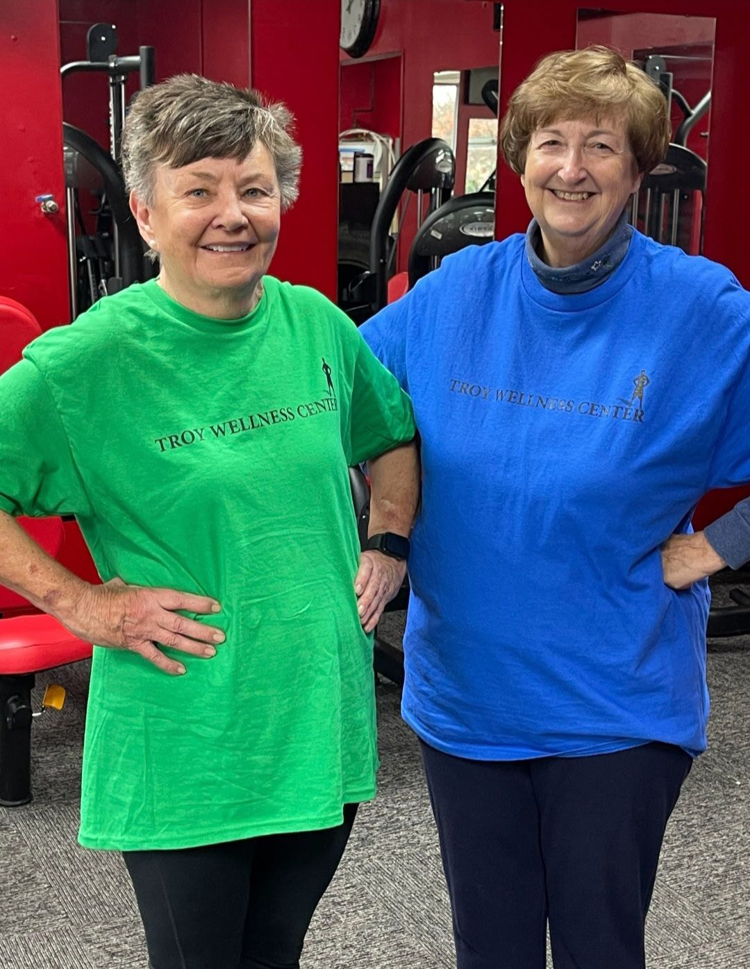 Two women smiling, posing at a gym, wearing matching shirts in green and blue, with hands on hips.