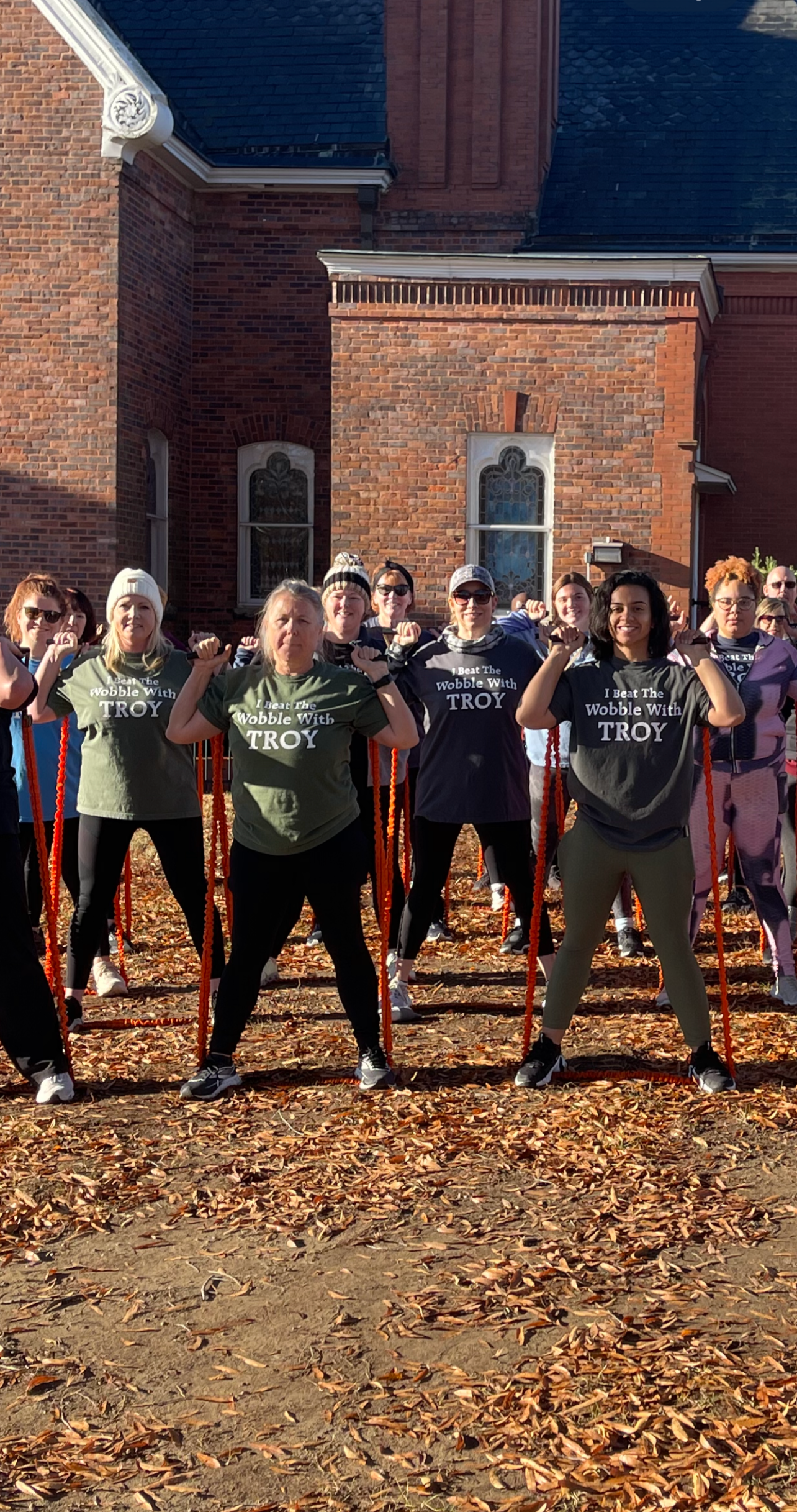 Group of women exercising outdoors near a brick building, autumn leaves on the ground.