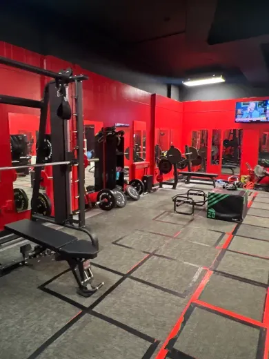 Red-walled gym with weight machines, free weights, and mirrors. Gray floor with black and red taped workout boxes.